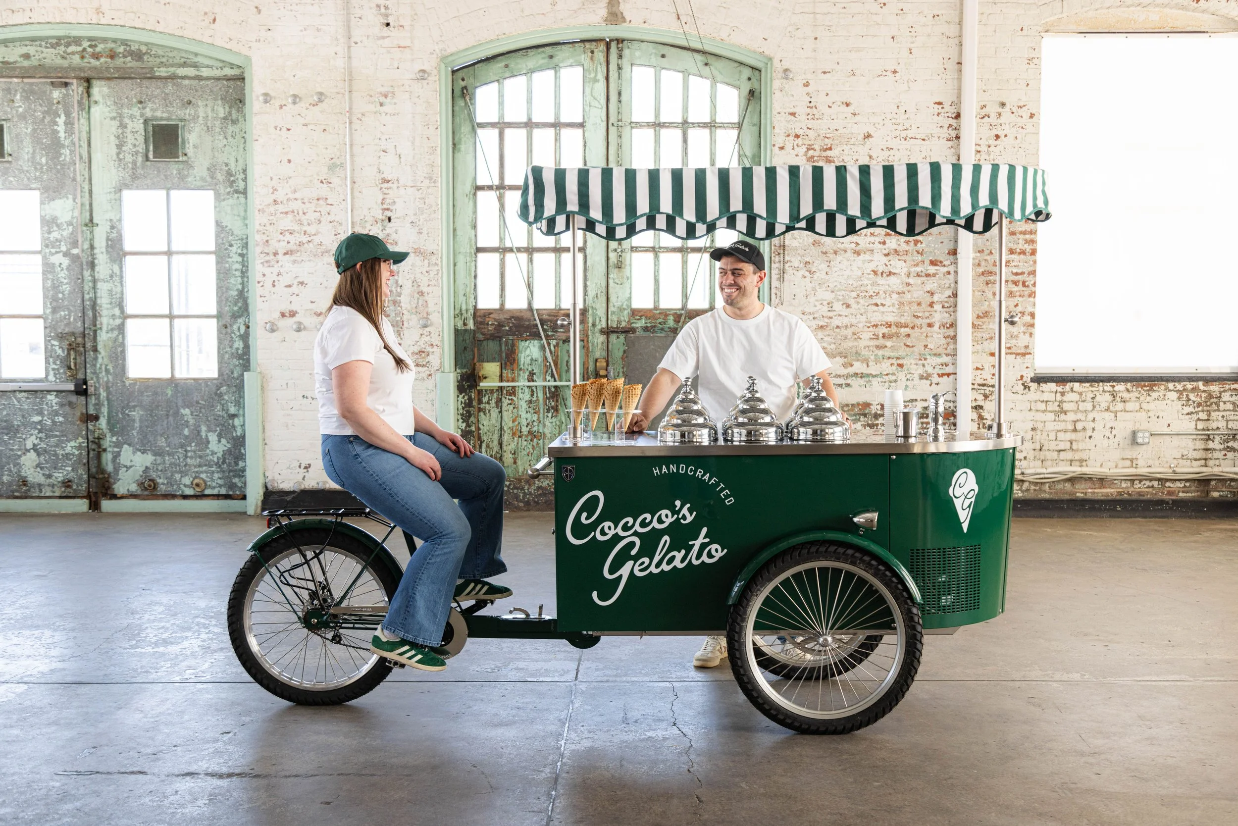Gelato cart titled 'Cocco's Gelato' with a woman sitting on the back of a bicycle, talking to a man operating the cart inside a rustic industrial space.