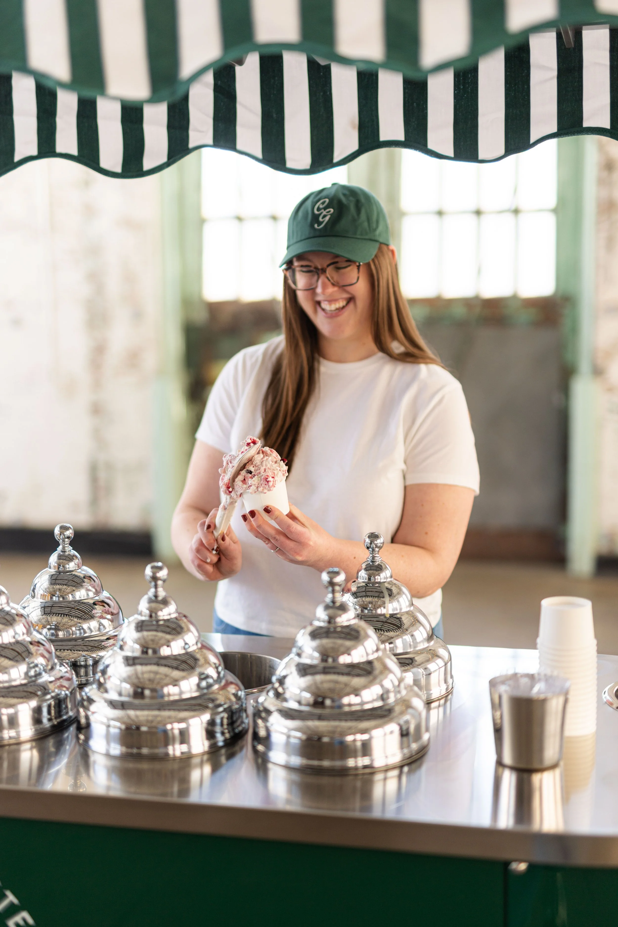 A woman (Jenica Cocco) smiling while holding a cup of gelato behind a gelato cart.