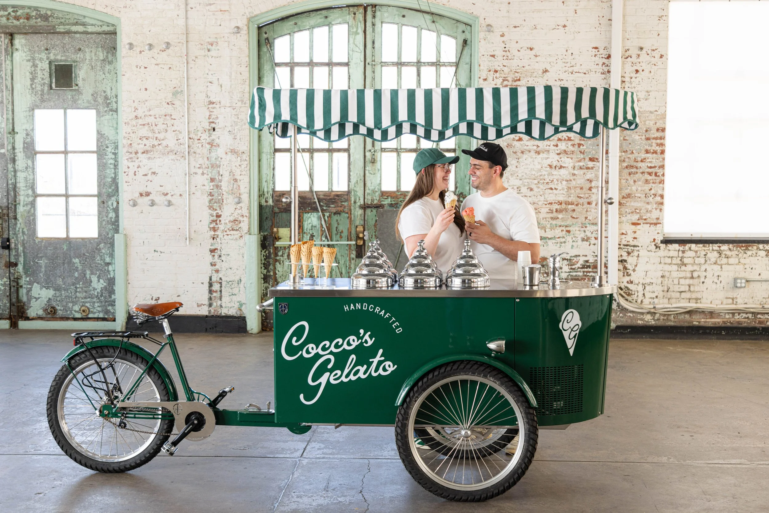 Two people stand behind a green gelato cart labeled 'Cocco's Gelato' in a warehouse setting. They are smiling and holding ice cream cones, sharing a moment together. The cart has a canopy and a bicycle attached to it.
