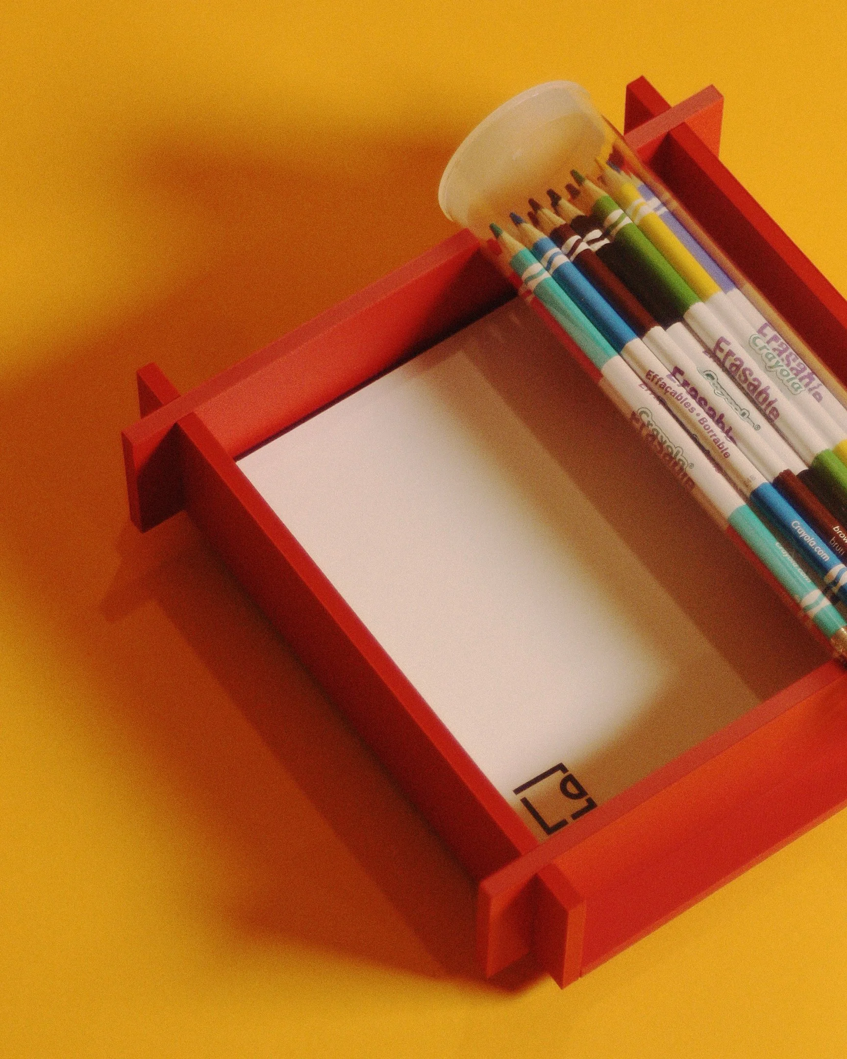 Red wooden tray containing a white notepad and a plastic case of colored pencils on a yellow surface.