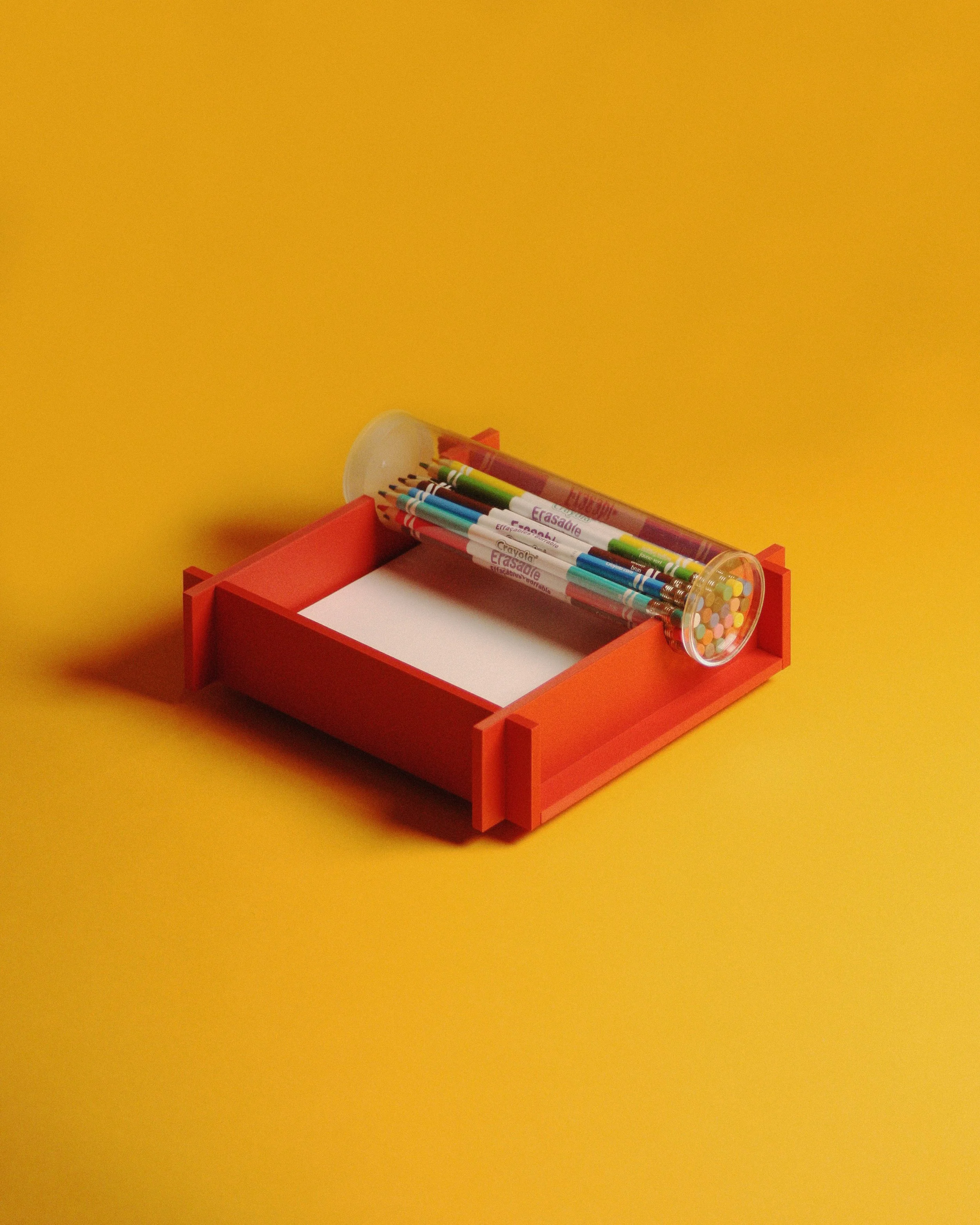 Orange storage box with a white notepad inside, topped with a tube of colorful gel pens and a container of multicolored confetti or beads, against a yellow background.