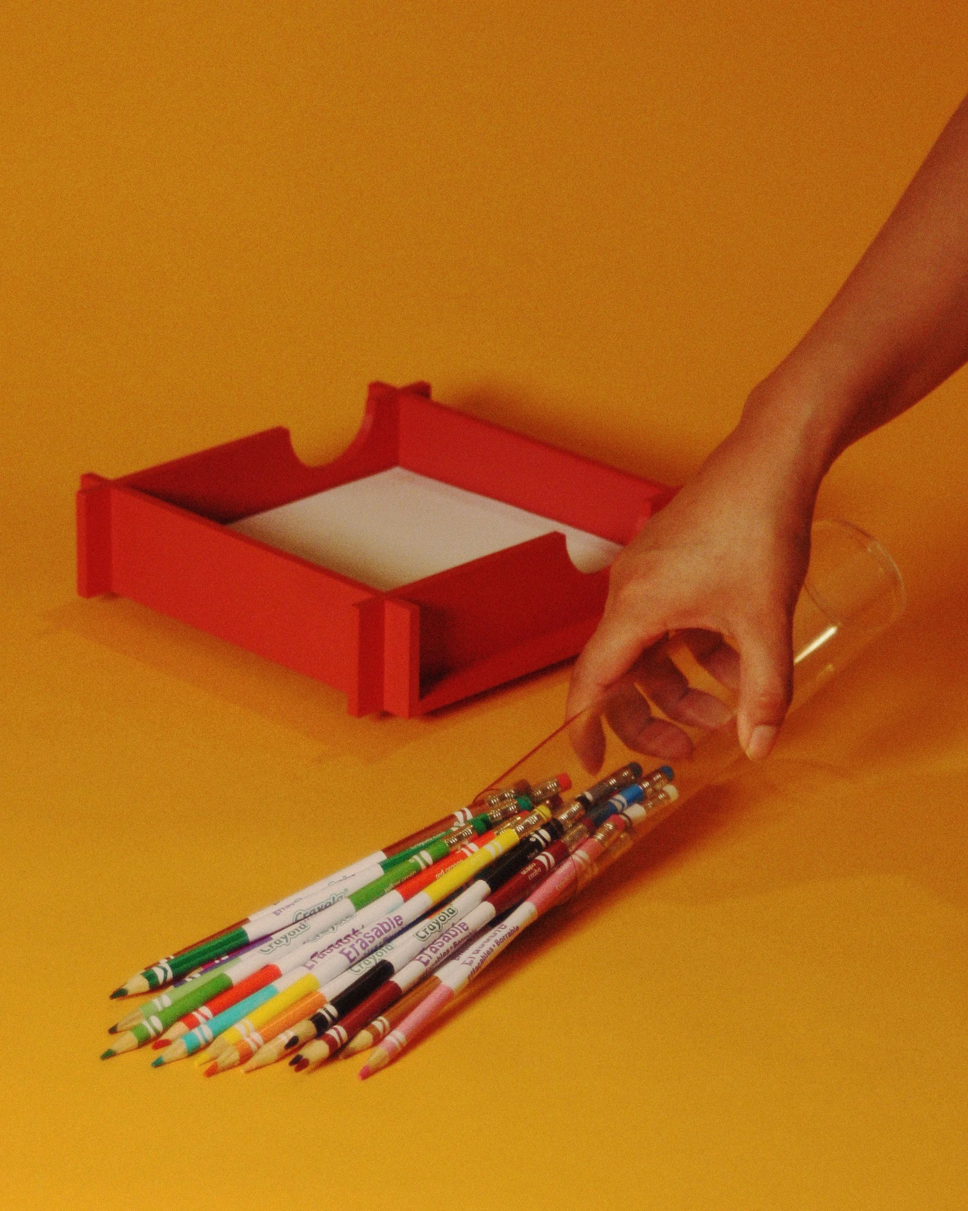 Hand holding a red and clear container filled with colorful Crayola markers, with a red pencil case in the background on a yellow surface.