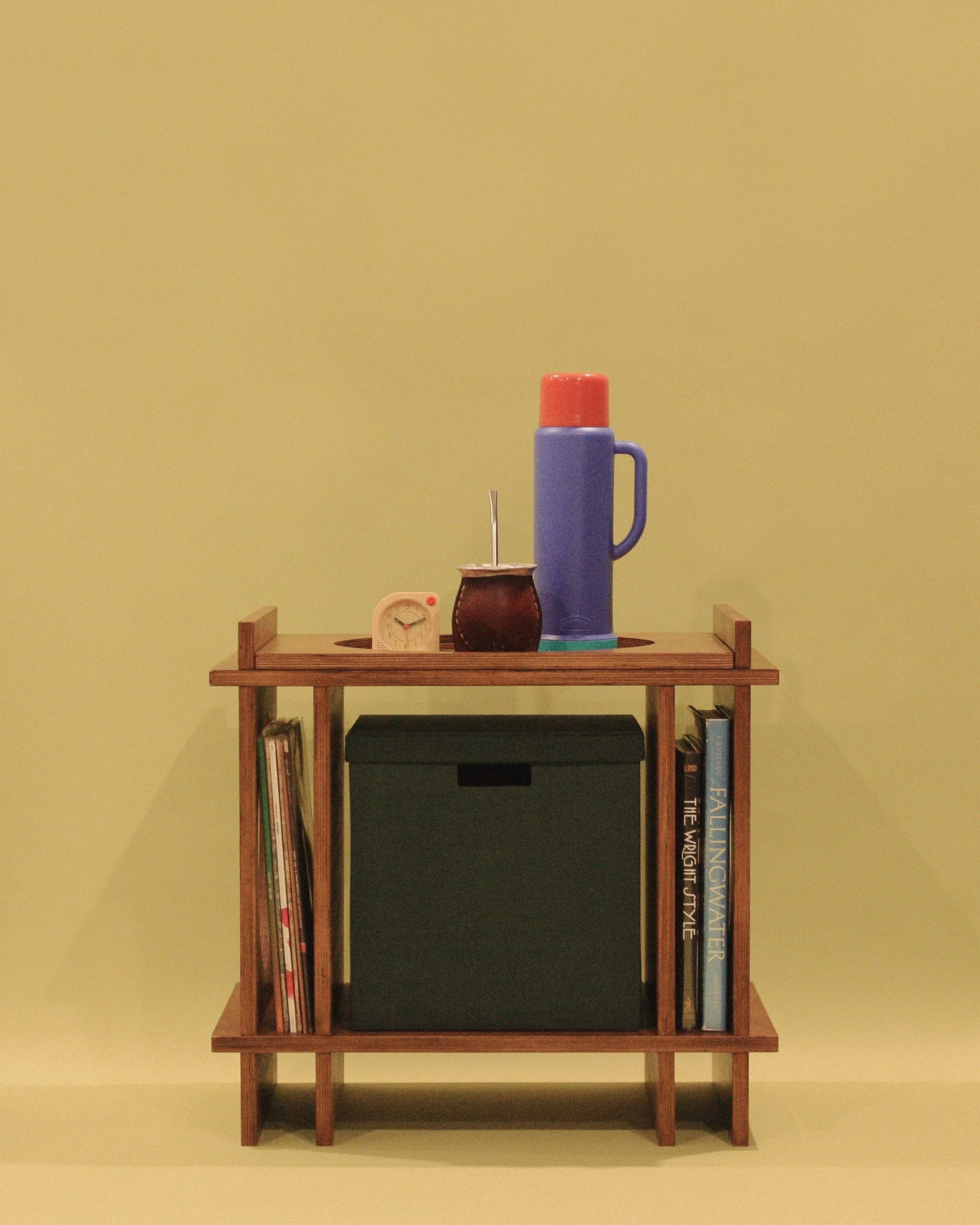 A wooden shelf against a yellow wall holding a small clock, a brown pot, a tall blue thermos with an orange lid, and books.