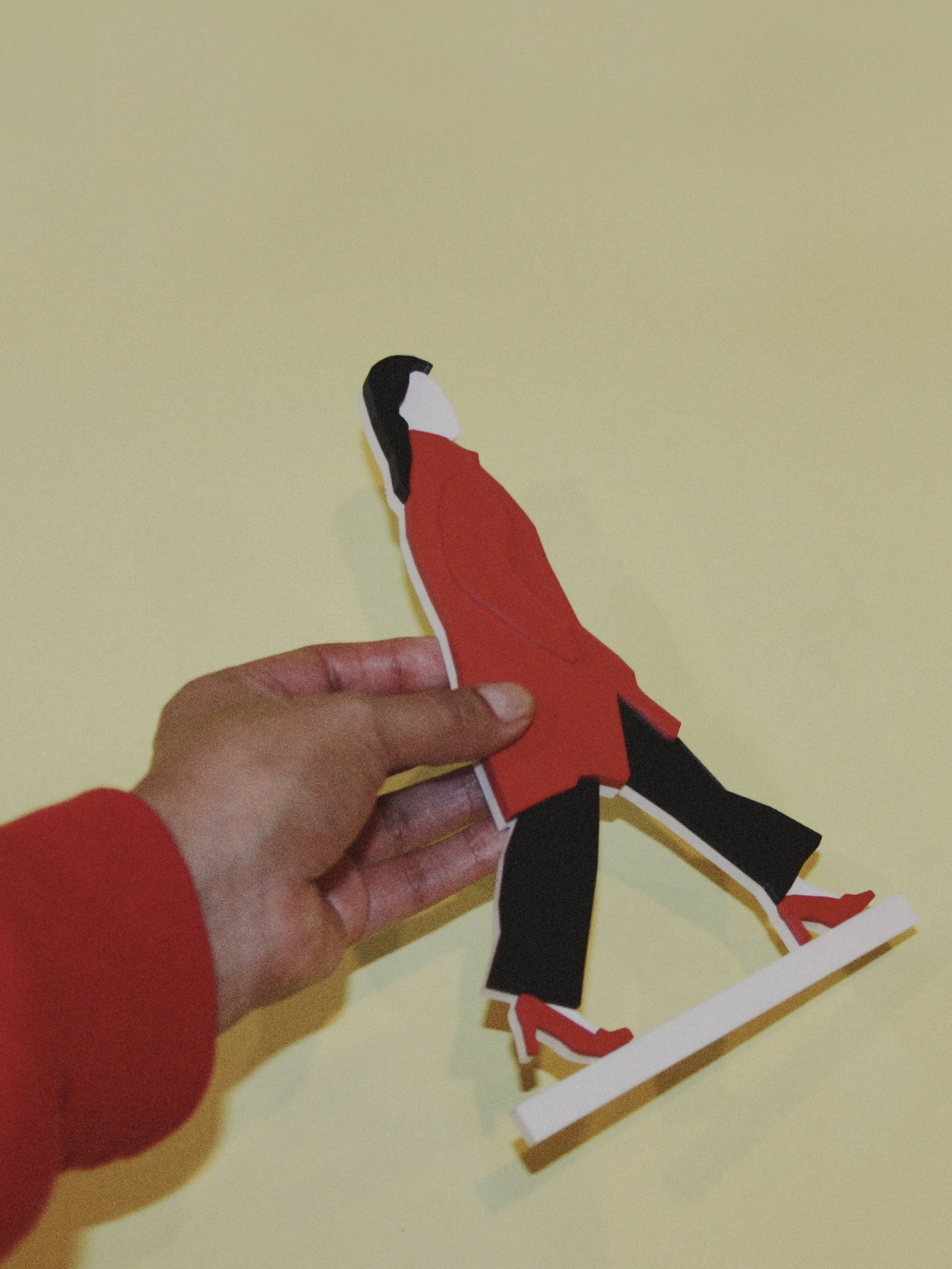 A hand holding a paper craft of a person ice skating, wearing a red jacket and black pants, against a yellow background.