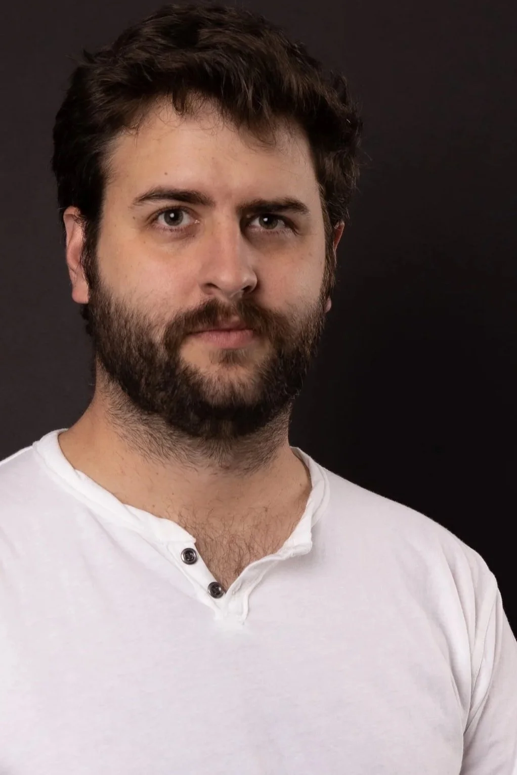 Close-up portrait of a man with dark hair and beard, wearing a white shirt, against a dark background.
