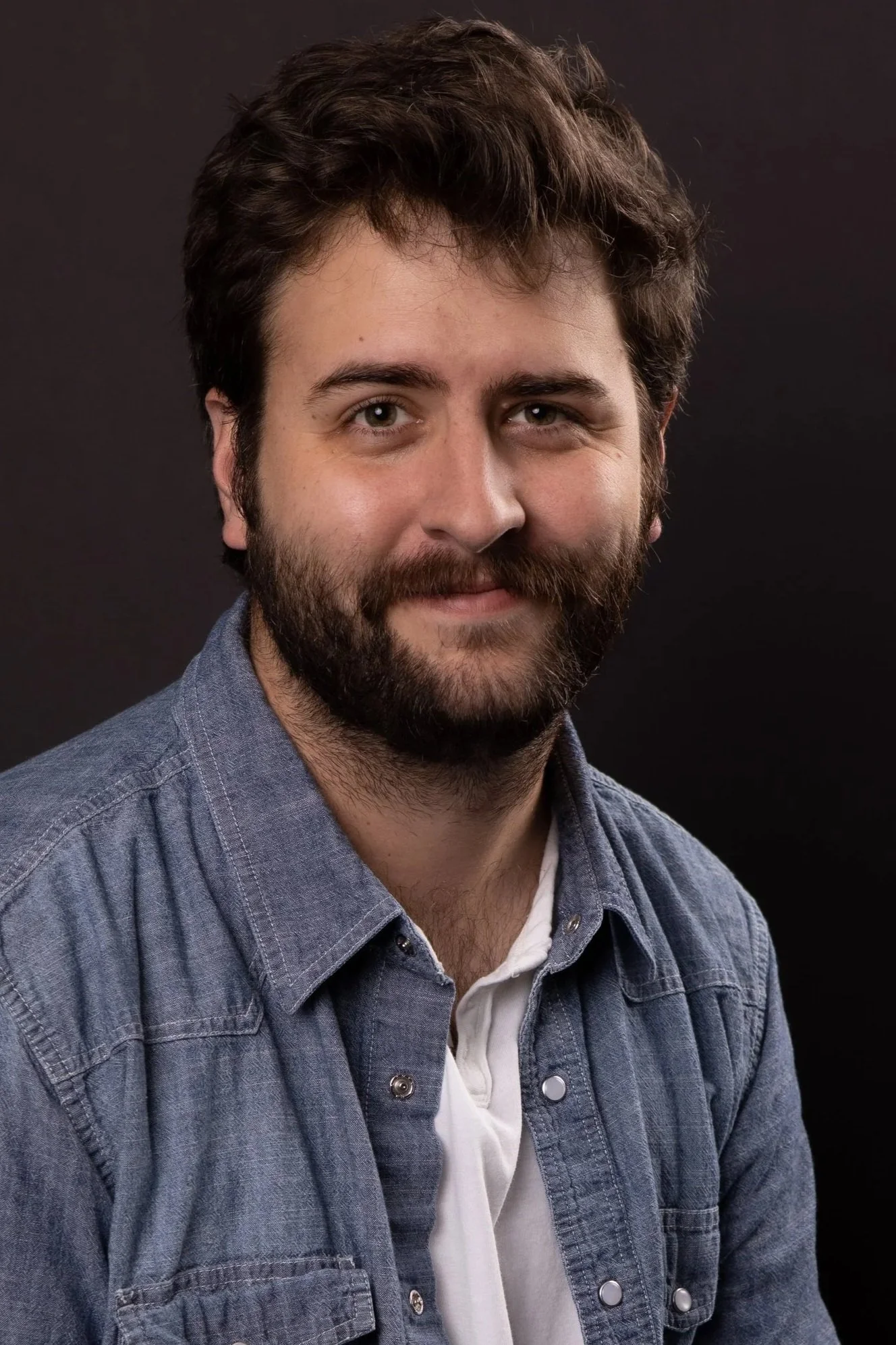 A man with dark, curly hair and a beard, wearing a denim shirt over a white T-shirt, smiling softly against a dark background.