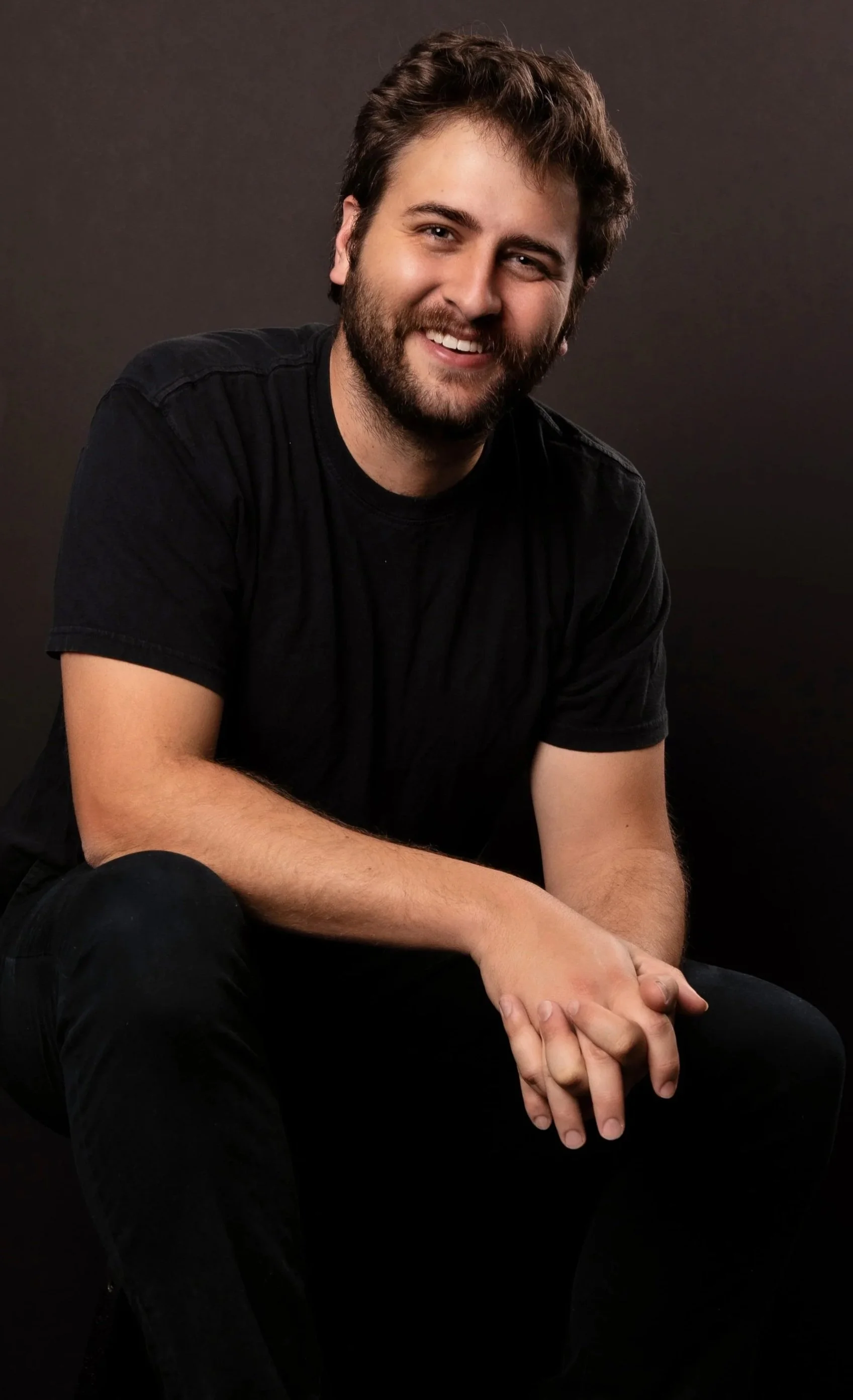A smiling young man with a beard and brown hair, wearing a black t-shirt, sitting against a dark background.