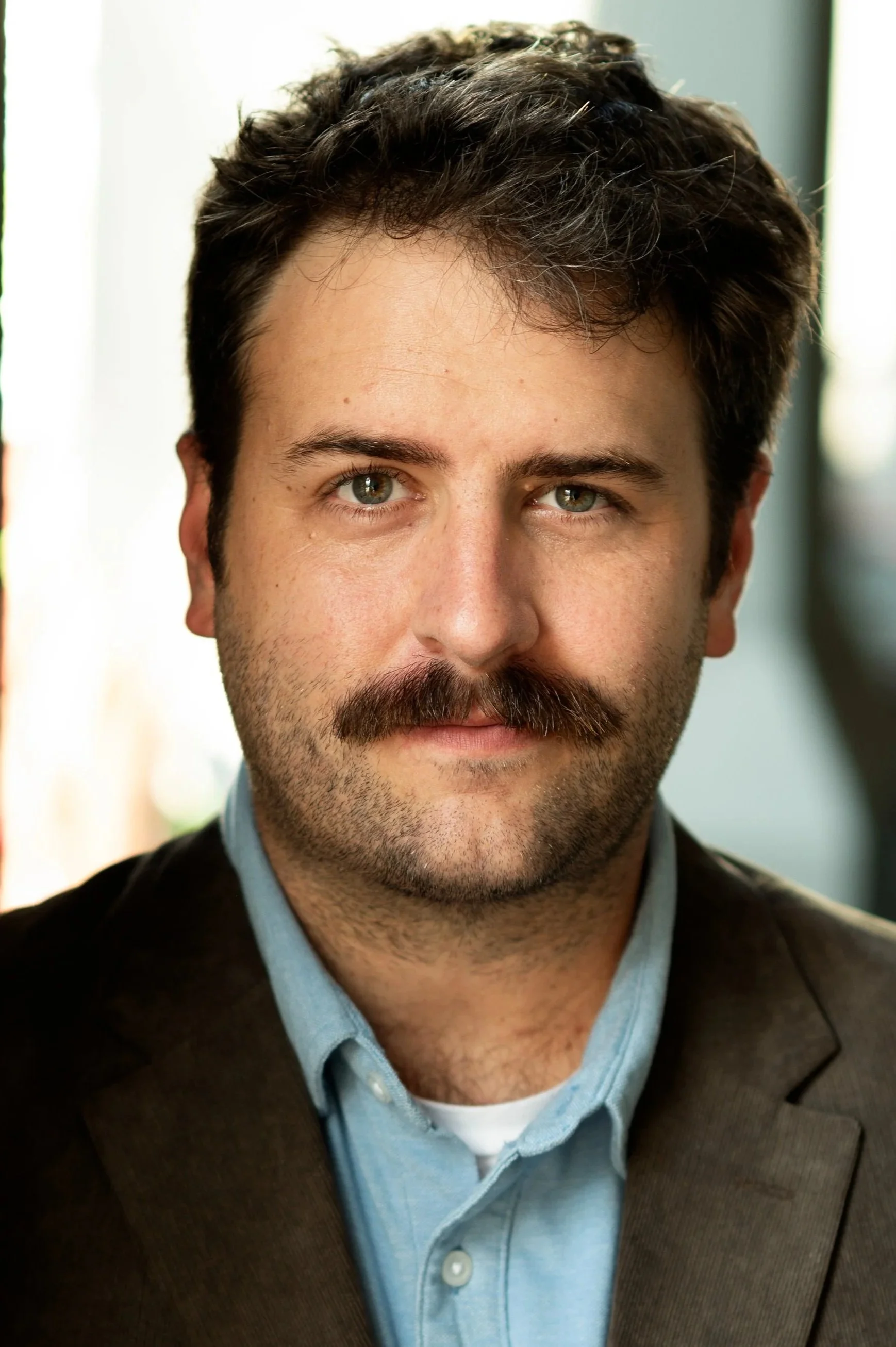 A man with dark, curly hair and a beard. He is wearing a blue collared shirt under a dark blazer. The background is blurred.