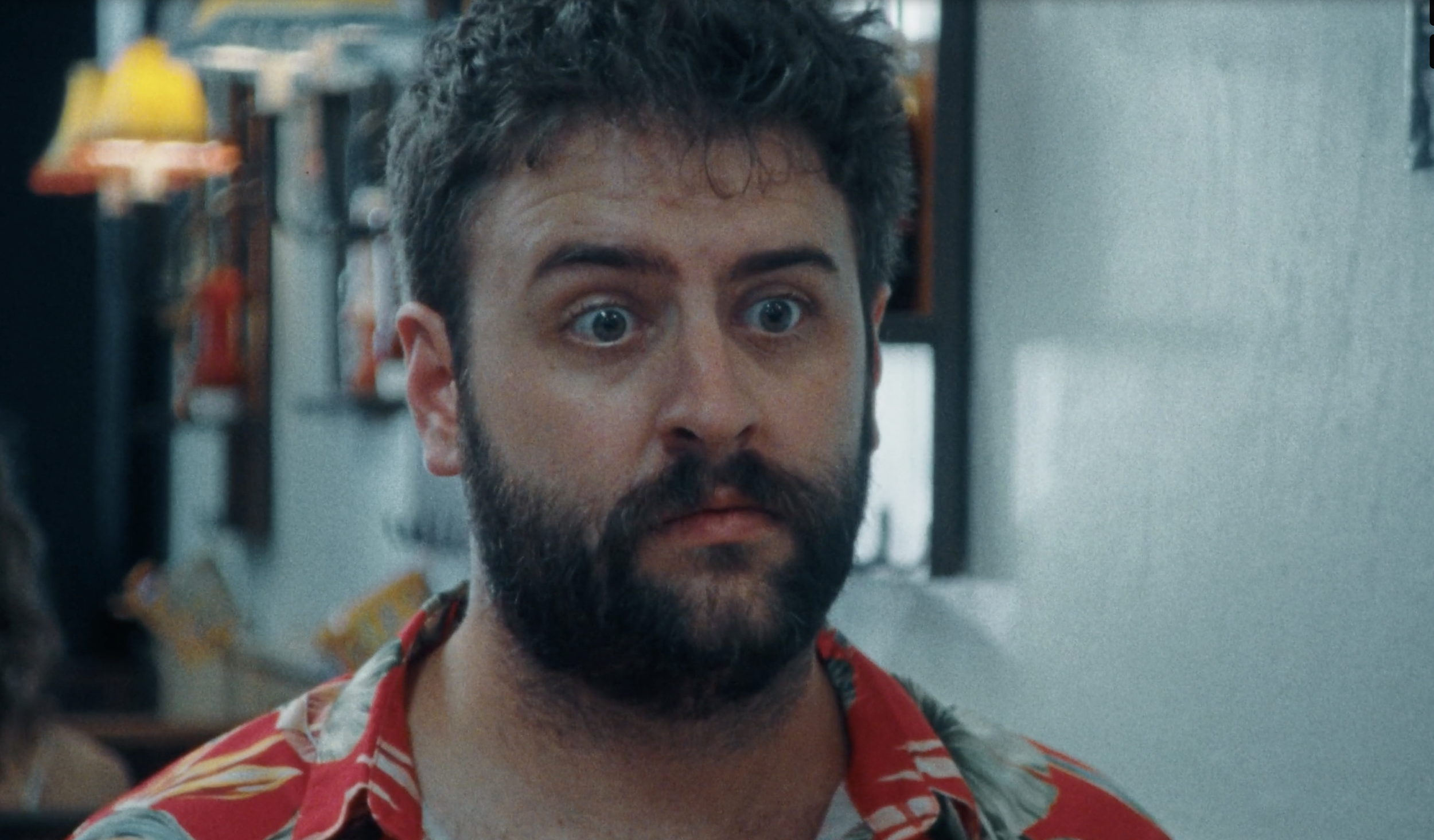 Close-up of a man with dark curly hair, blue eyes, and a beard, wearing a red floral shirt, in a store or bakery with blurred shelves and baking supplies in the background.