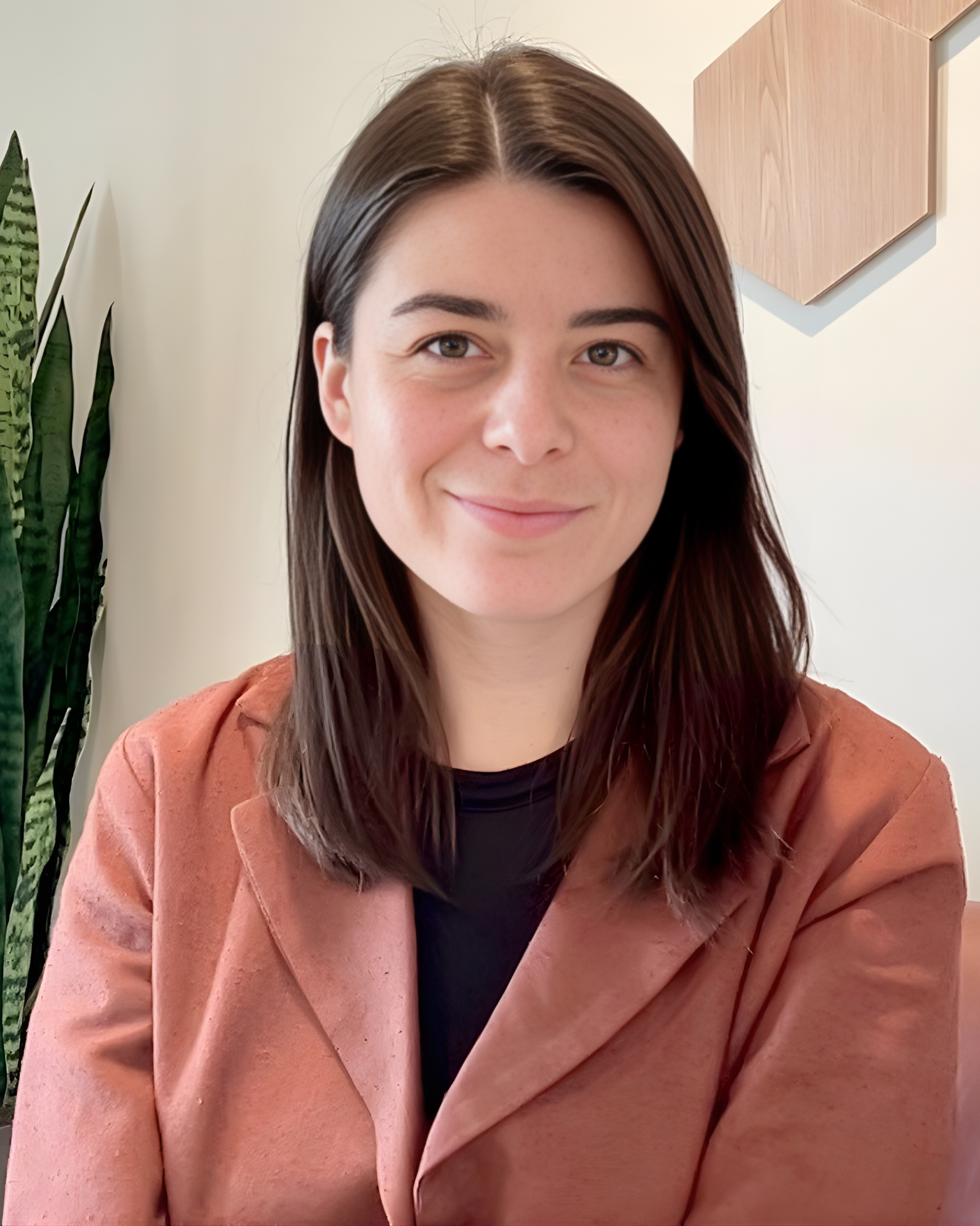 A young woman with shoulder-length brown hair smiling indoors, wearing a salmon-colored blazer and black top, with a tall green plant and light-colored wall with wooden wall art in the background.