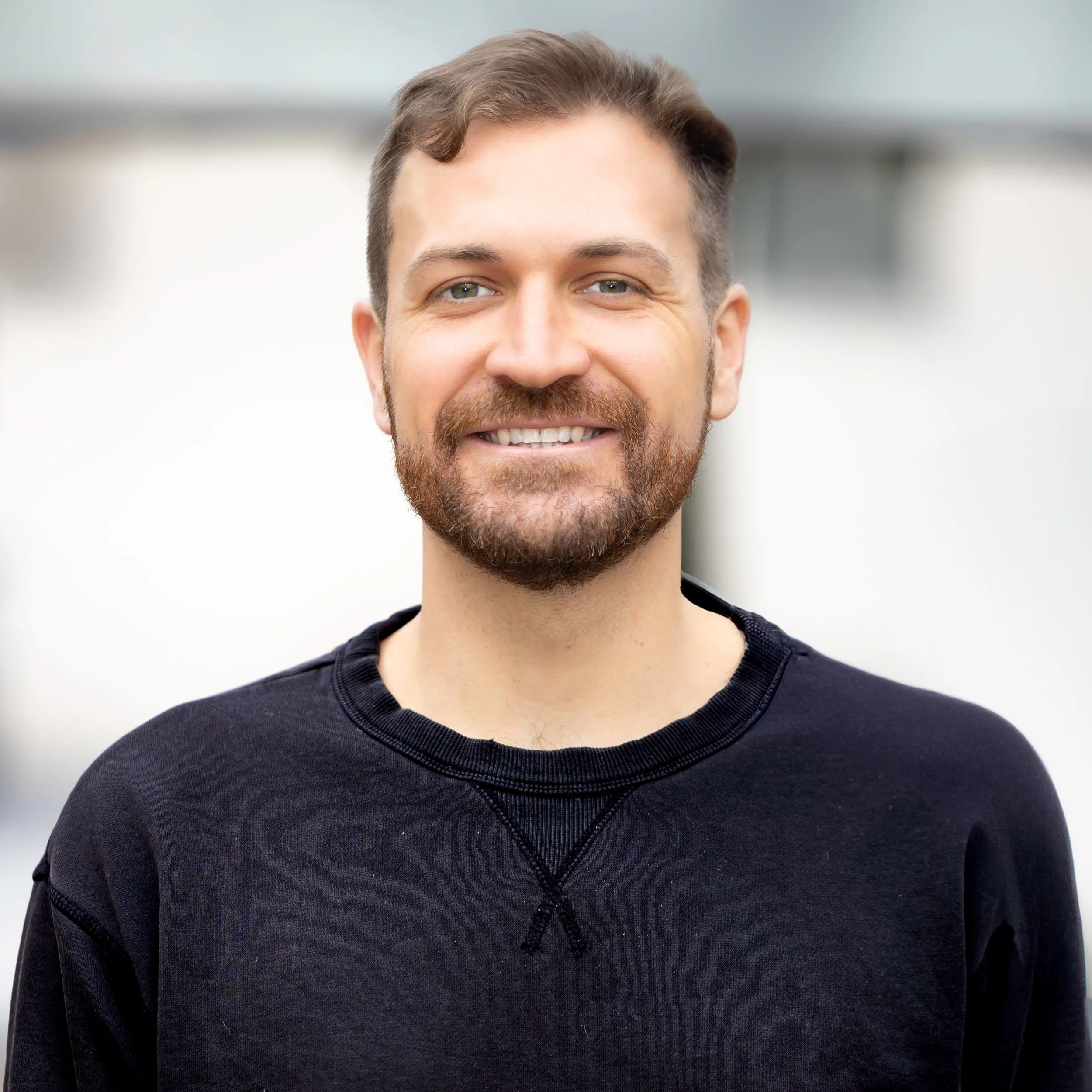 Close-up of a smiling man with a beard and short hair, wearing a black sweatshirt, outdoors in daylight.