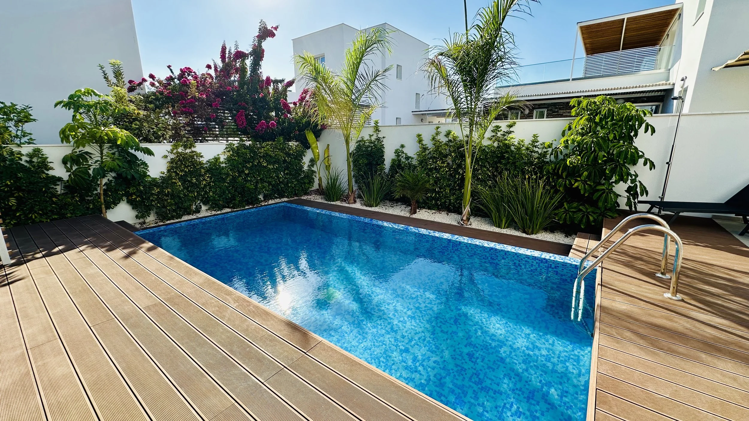 Outdoor swimming pool with wooden deck, lush green plants, and a modern house in the background. Spacious family villa in Paphos with a private pool.