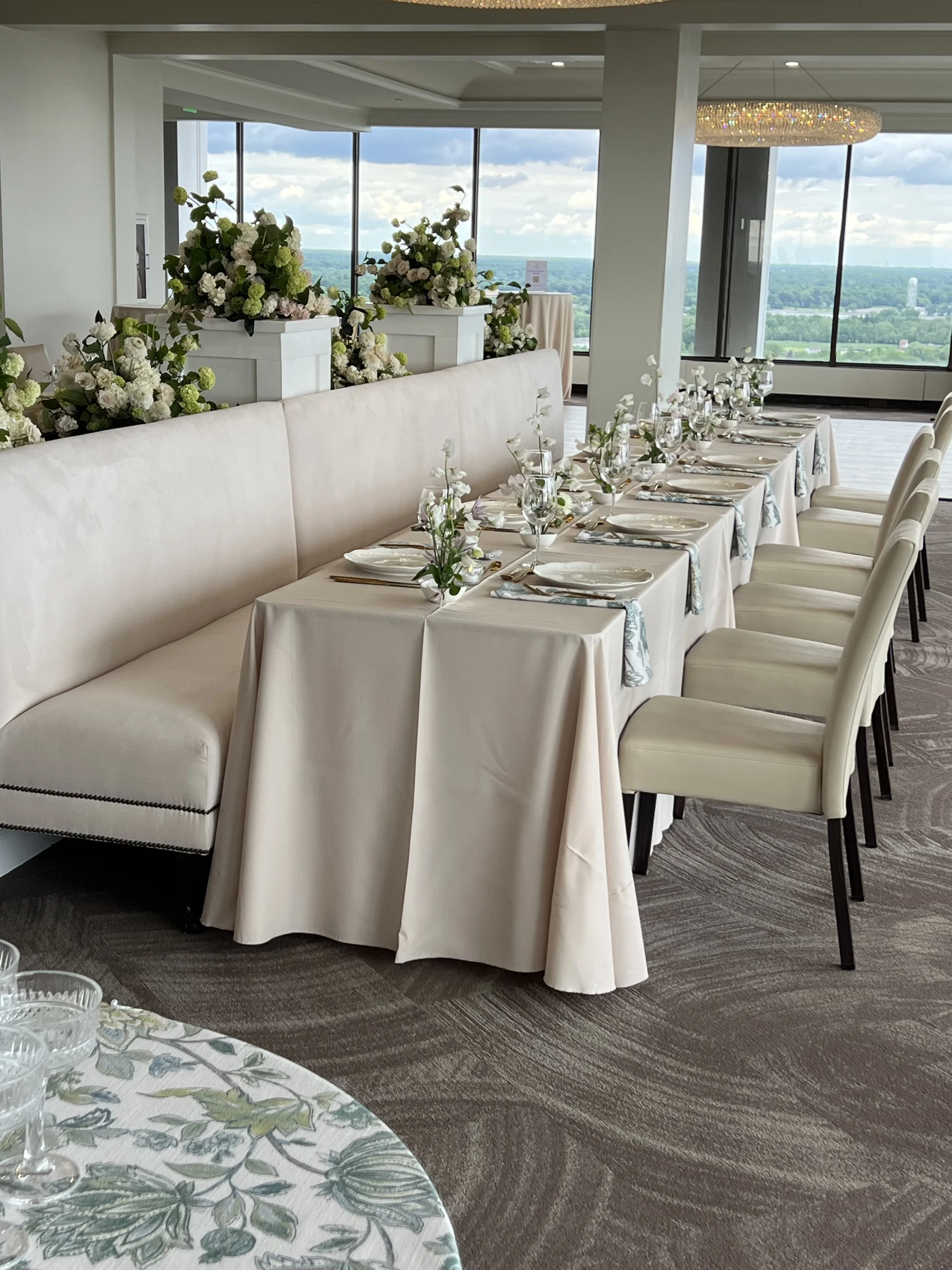 Round tables covered in white tablecloths, each set with glassware, plates, and napkins. Tall floral centerpieces with white and pink flowers decorate the tables. Large windows offer a city view, and modern chandeliers hang from the ceiling.