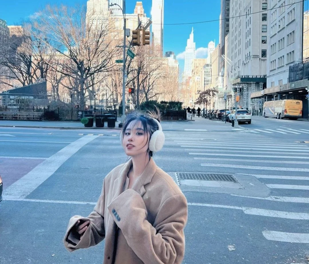 A young woman wearing earmuffs and a beige coat holding a phone stands at a crosswalk in an urban cityscape, with tall buildings, cars, and a partly cloudy sky in the background.