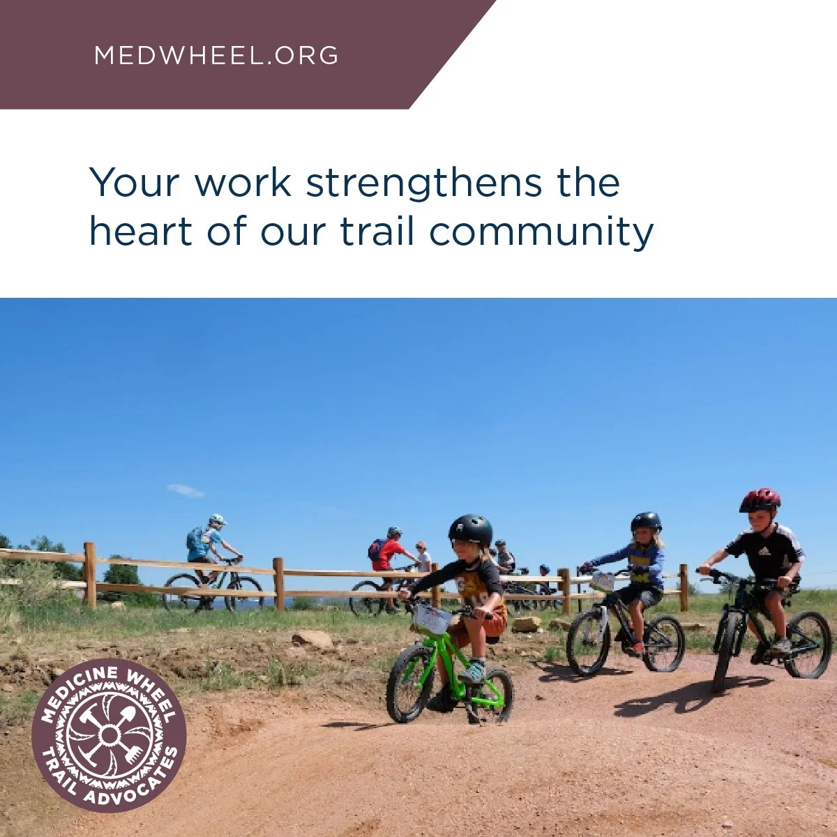 Children riding bikes on a dirt trail with a clear blue sky, wooden fence, and more cyclists in the background, promoting trail advocacy.