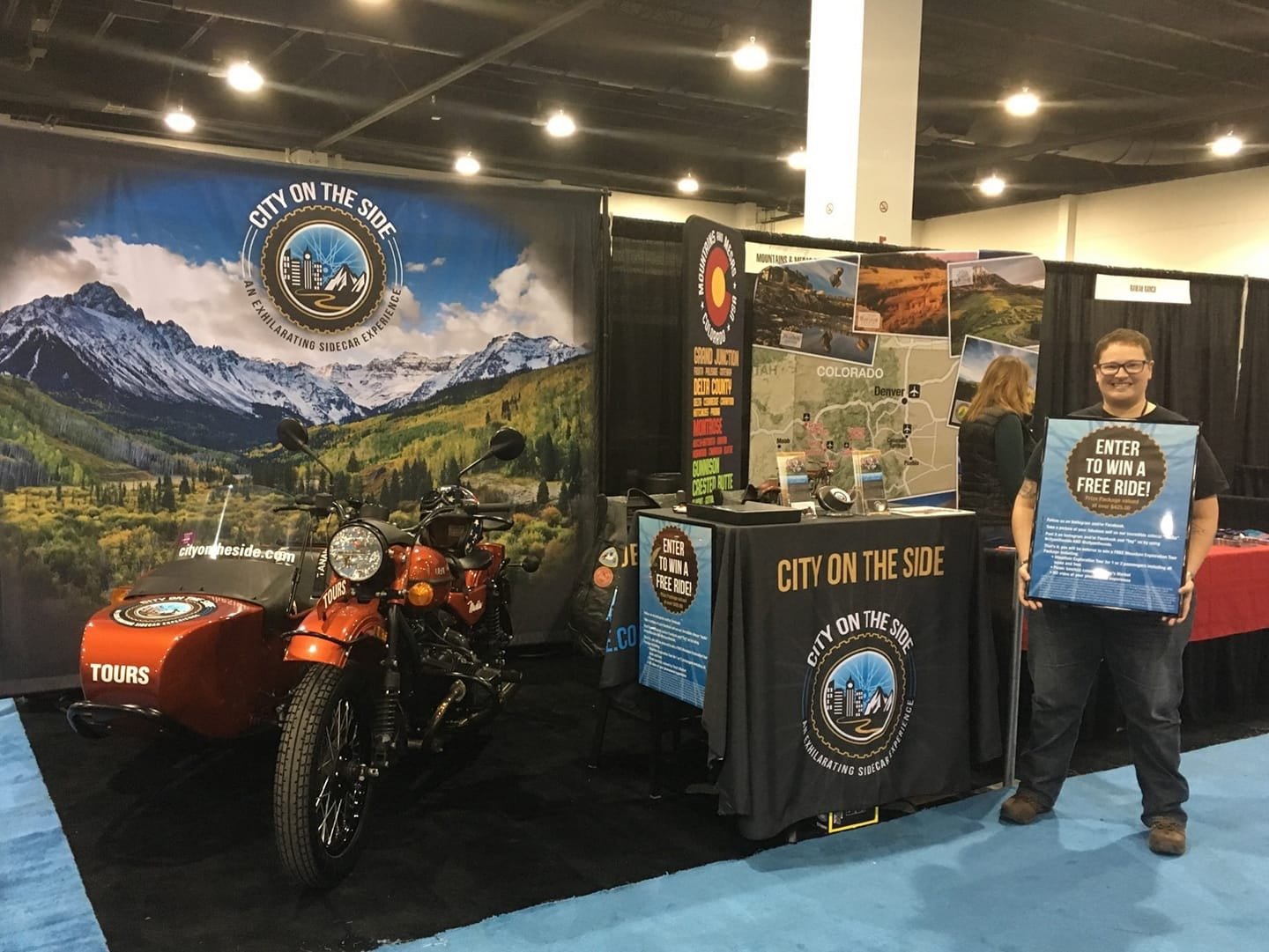 Tradeshow booth with a mountain-themed backdrop and a motorcycle with a sidecar, promoting cityside tours in Colorado. A man holds a sign advertising a contest to win a free ride. The booth features a table with brochures and informational materials.