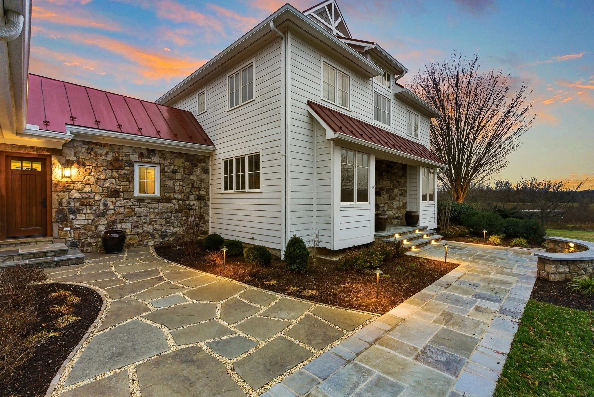 Exterior view of a modern two-story house with a stone and white siding facade, a red metal roof, a stone pathway with landscape lighting, and a landscaped yard with trees at sunset.