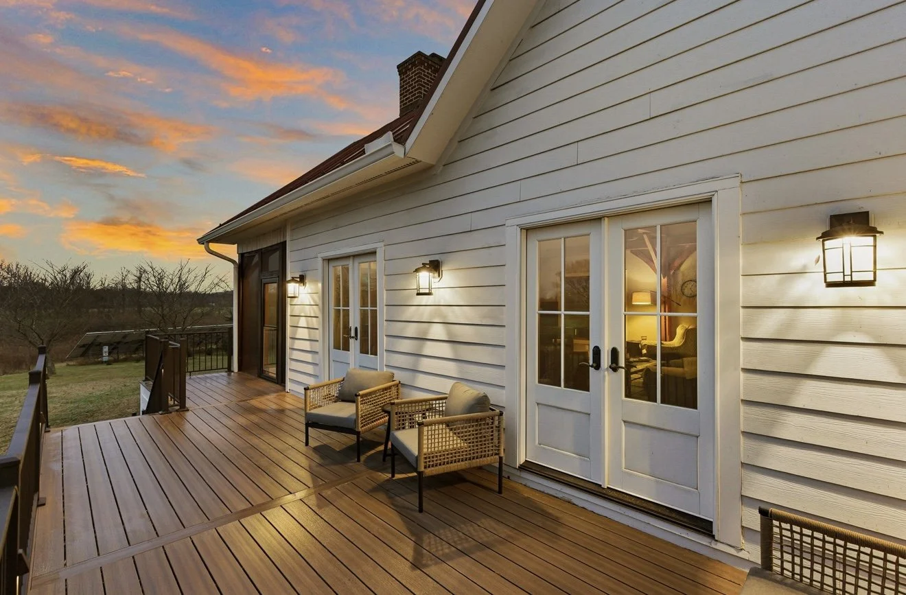 A wooden deck attached to a white house with double glass doors, outdoor wall lights, and two chairs, overlooking a sunset sky and a landscape with trees.