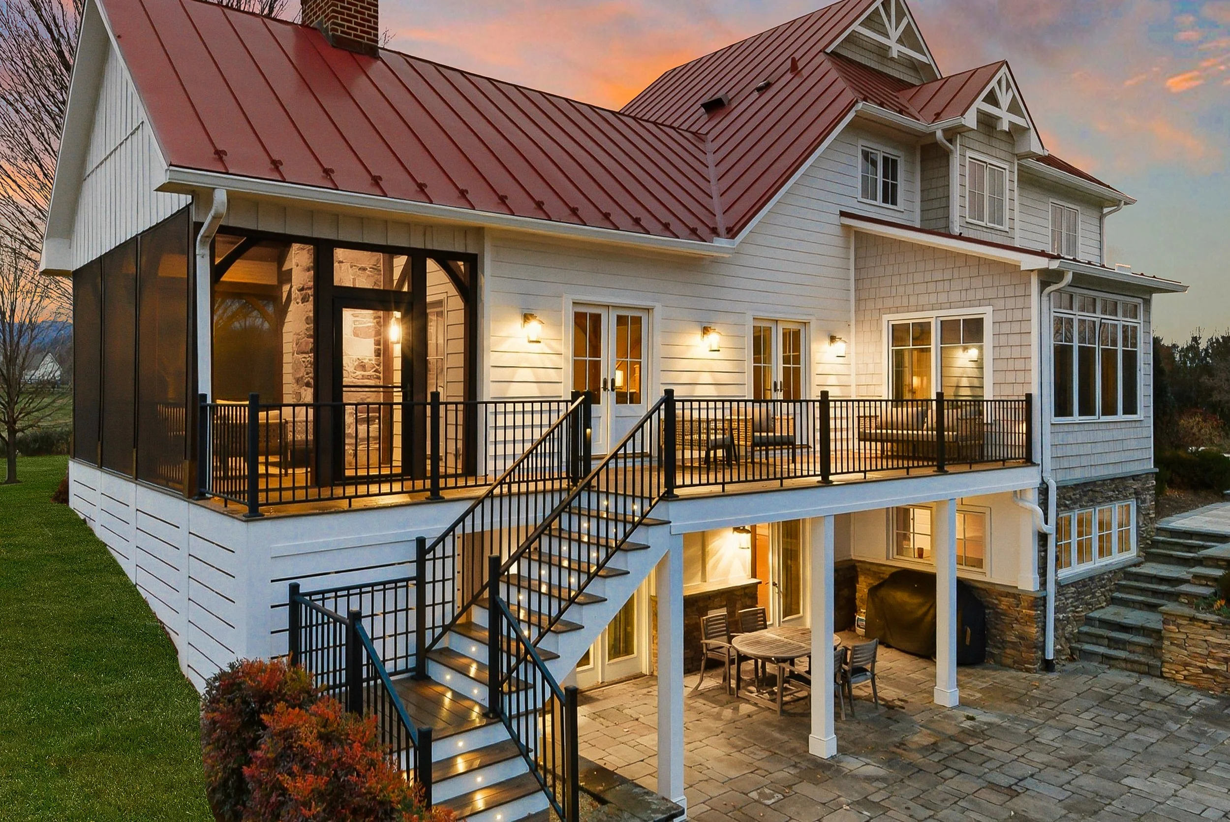 A multi-story house with white siding and stone accents, red metal roof, and a large porch lit by exterior lights at sunset.