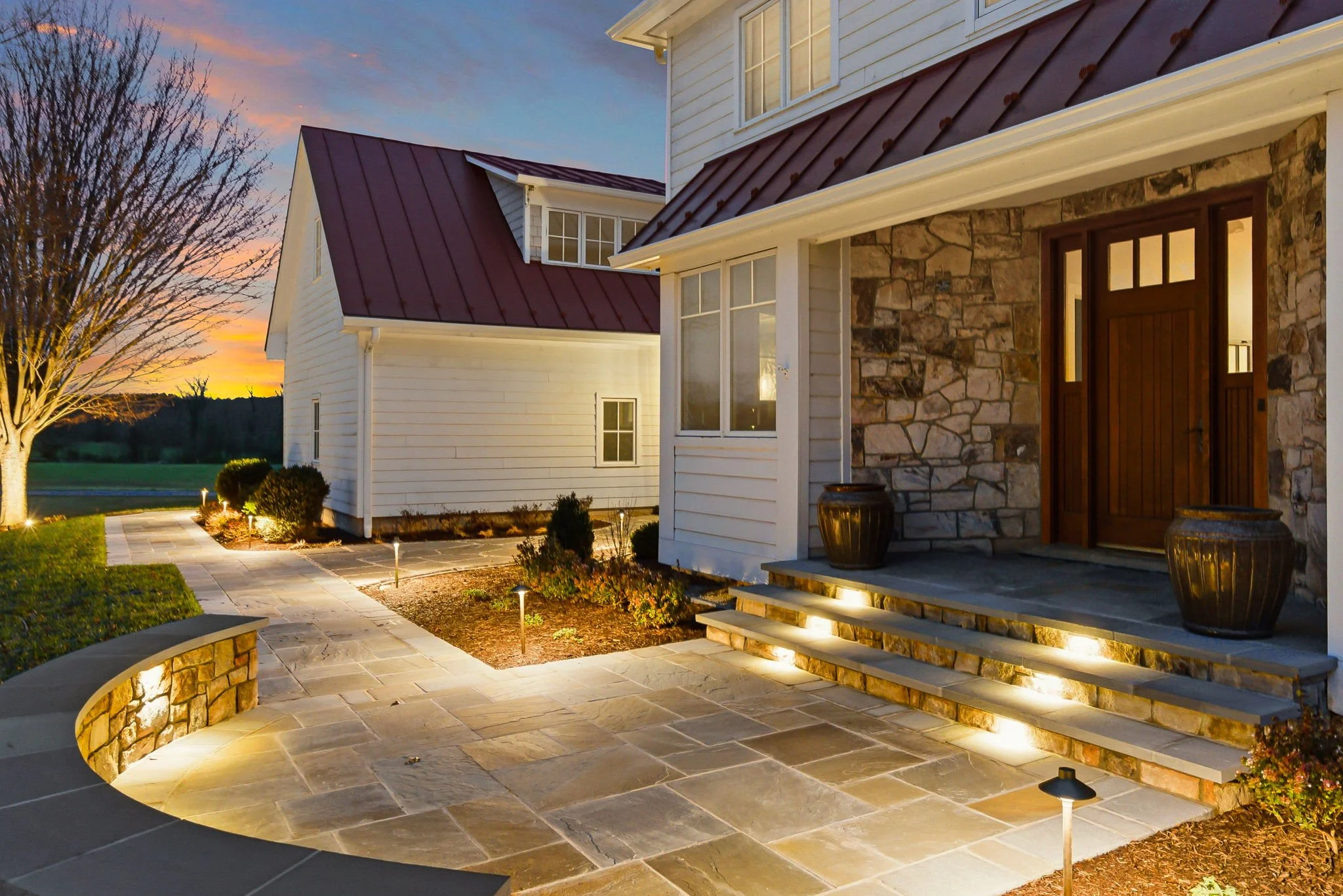 A front porch area of a house with steps, large potted plants, and pathway lights, during sunset.