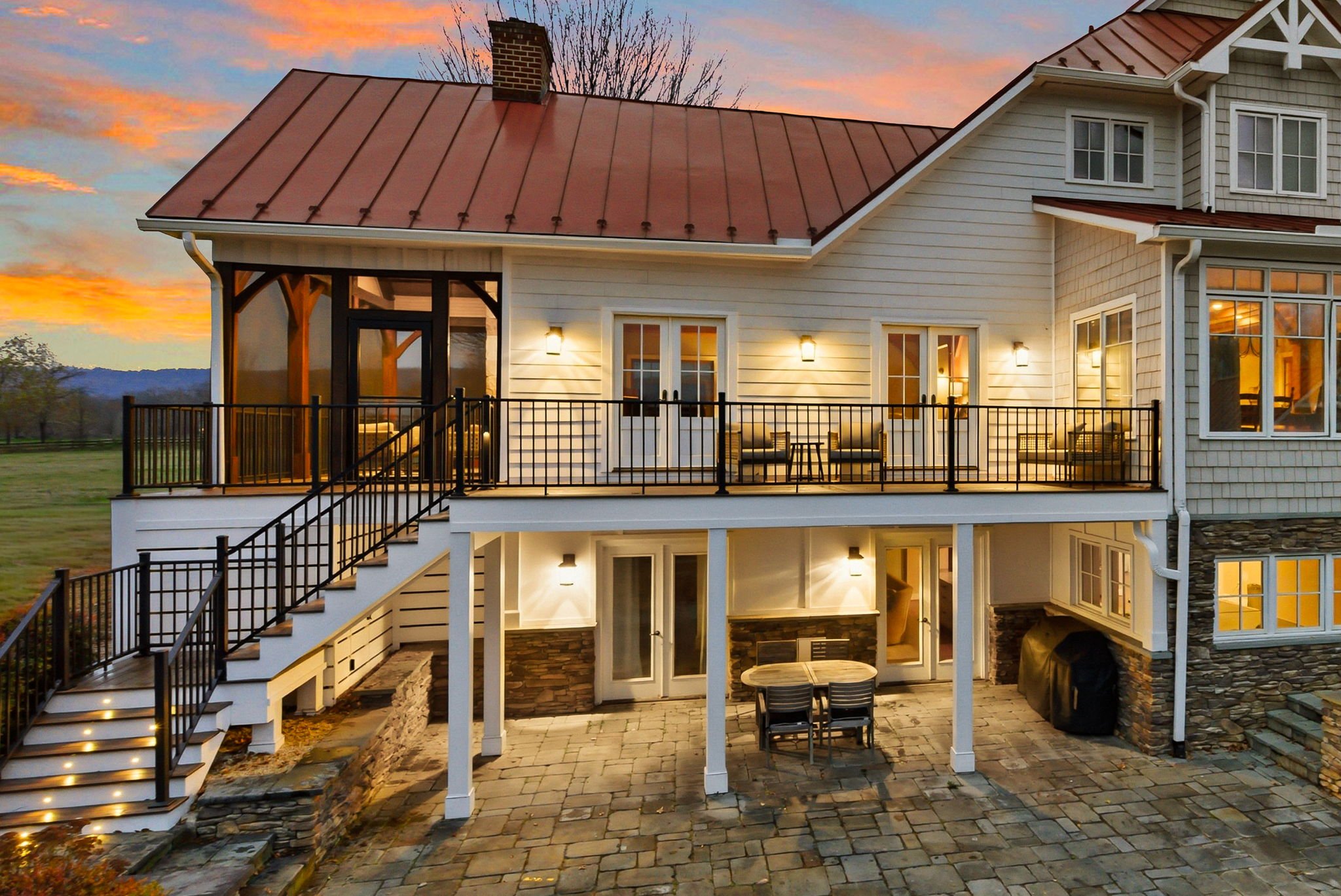 A multi-story house with a red metal roof, white siding, and a stone foundation. It has a large upper balcony with black railings and outdoor seating, and a lower patio with a table and chairs. The house is lit with exterior lights, and the sky in th