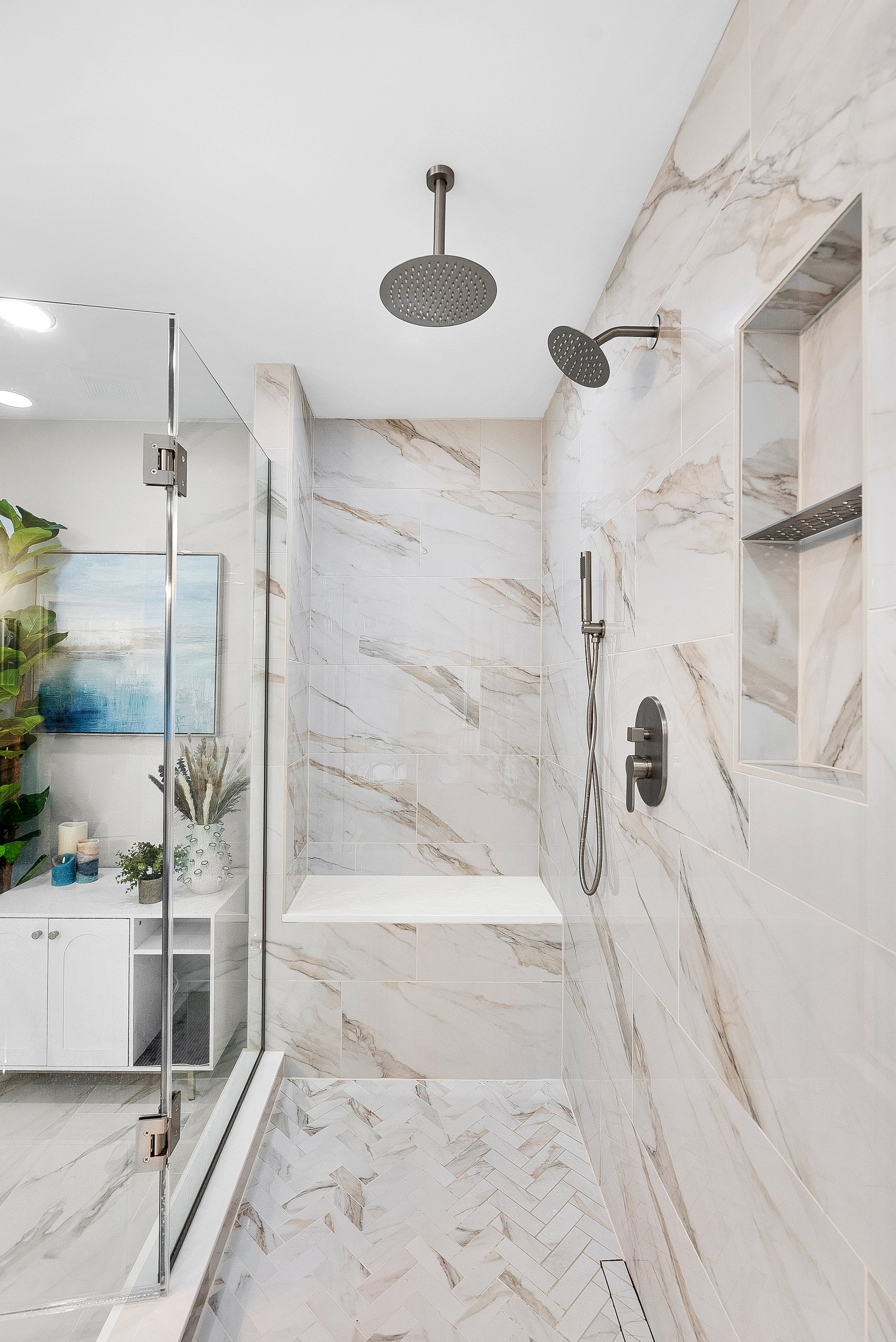 Modern bathroom shower with marble walls, a rain showerhead, a handheld shower, built-in shelf, and glass enclosure.