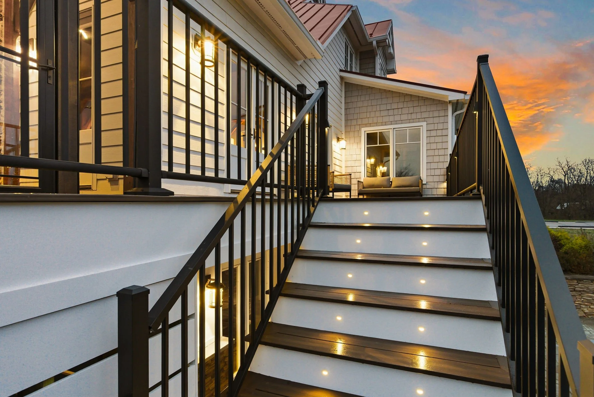 Exterior view of a house's upper porch and staircase at sunset, featuring black metal railings, outdoor lighting, and seating area.