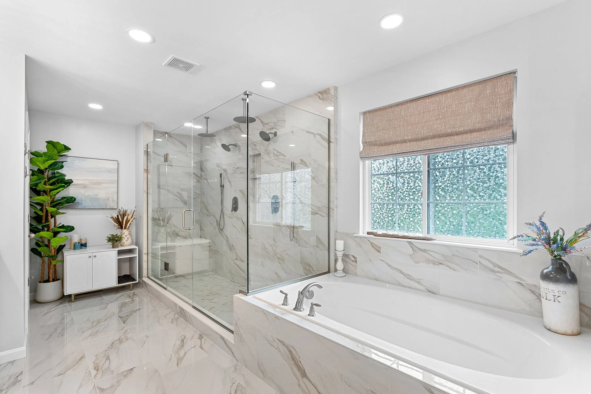 Modern bathroom with walk-in shower with glass walls, bathtub, window with beige shade, potted plant, and decorative items.
