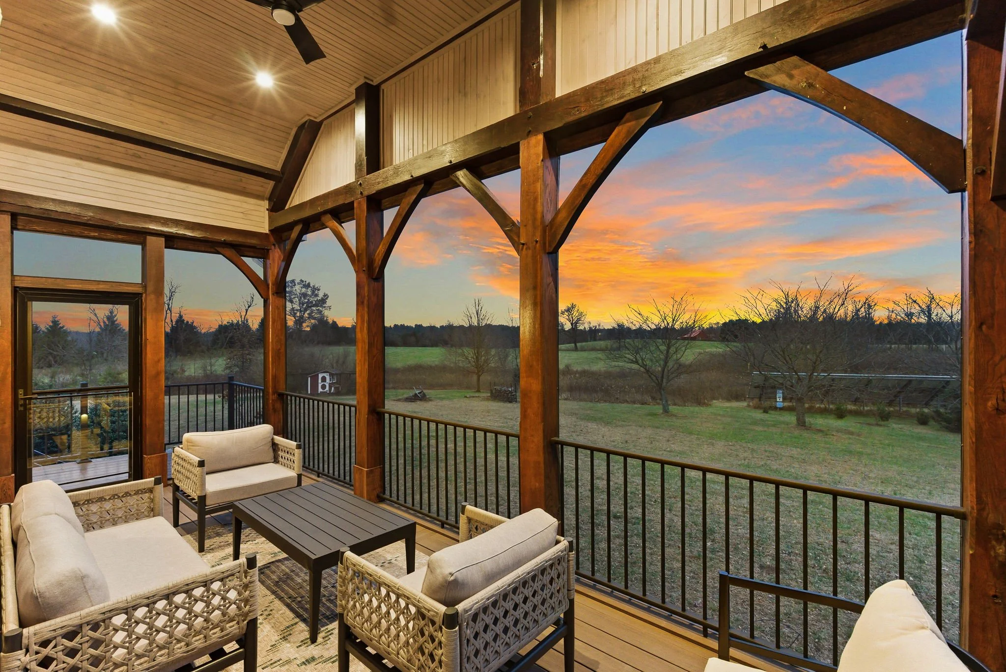 View from a screened porch with wooden beams, outdoor seating with cushions, overlooking a field and trees at sunset