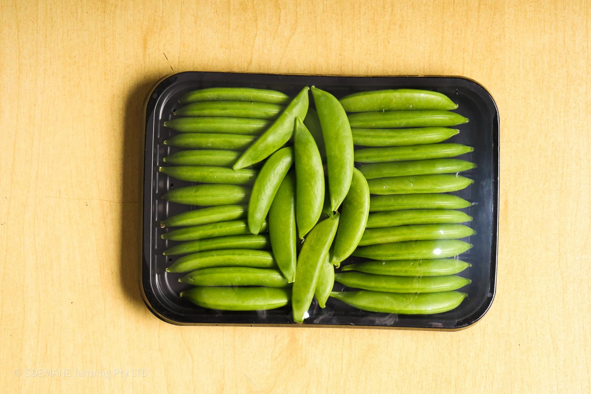 Tray of fresh green sugar snap peas on a wooden surface.