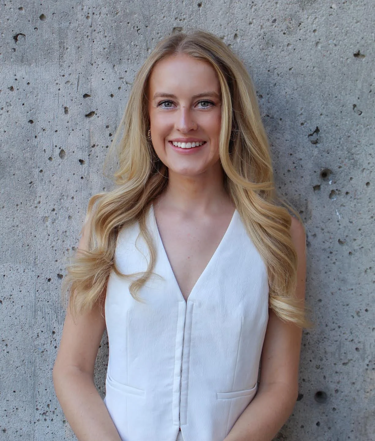 A young woman with long blonde hair wearing a sleeveless white top, smiling in front of a textured gray concrete wall.