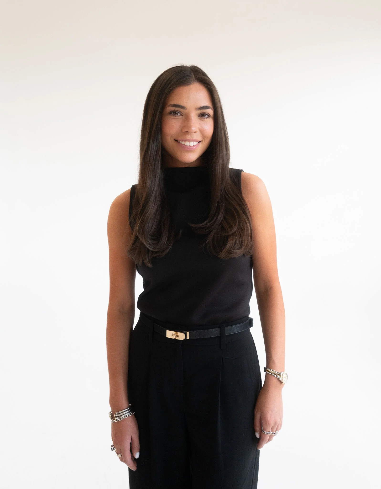 A woman with long dark hair wearing a sleeveless black top and black pants, accessorized with bracelets, rings, and a watch, smiling against a plain white background.