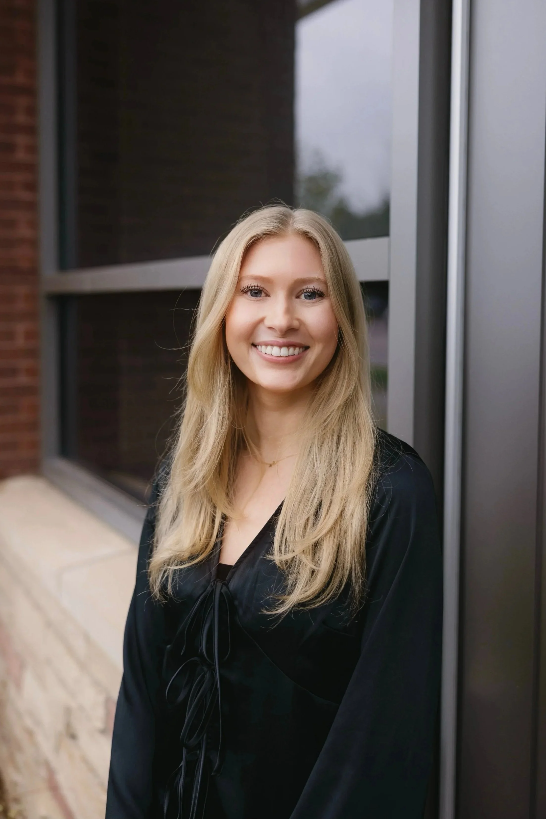 A young woman with long blonde hair, smiling, standing outdoors near a building with large glass windows, dressed in a black top.