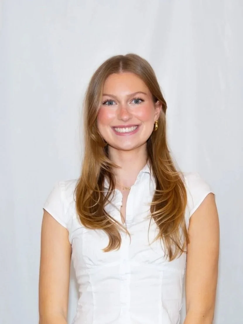 A young woman with long red hair, bright blue eyes, and a wide smile, wearing a white sleeveless blouse and gold hoop earrings, standing against a plain white background.