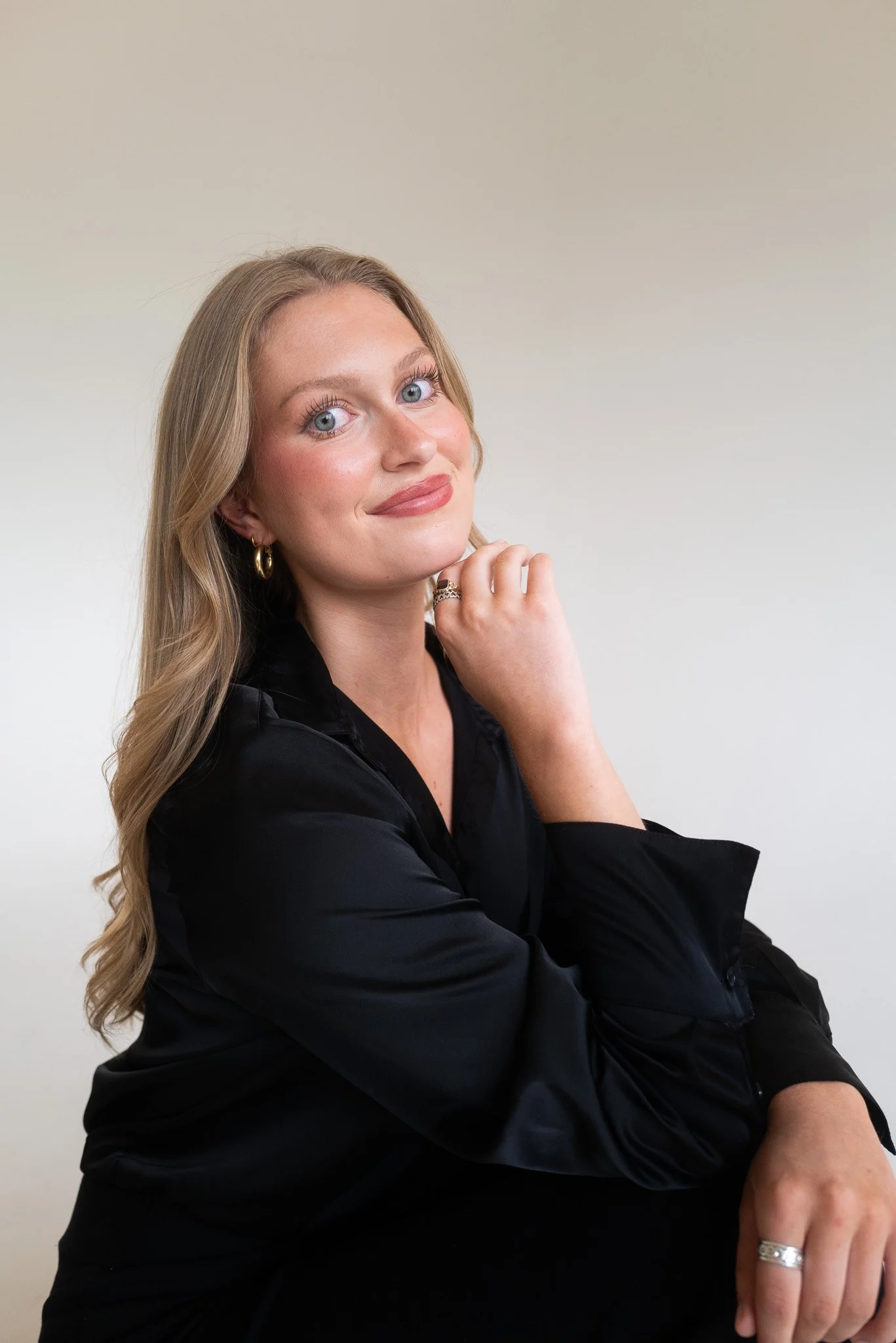 A woman with blonde hair, blue eyes, and light makeup, wearing a black shirt, gold hoop earrings, and rings, posing with her hand near her chin against a plain light-colored background.