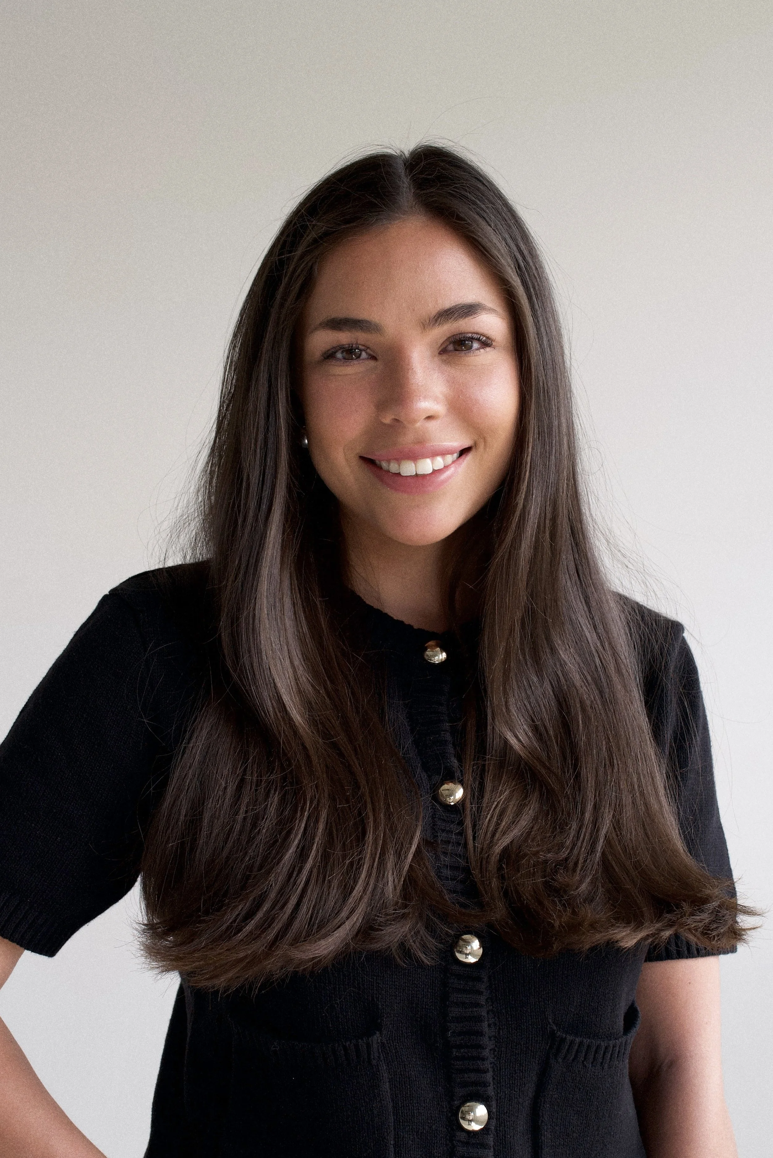 A young woman with long brown hair, smiling, wearing a black short-sleeved cardigan with large gold buttons, against a plain light background.