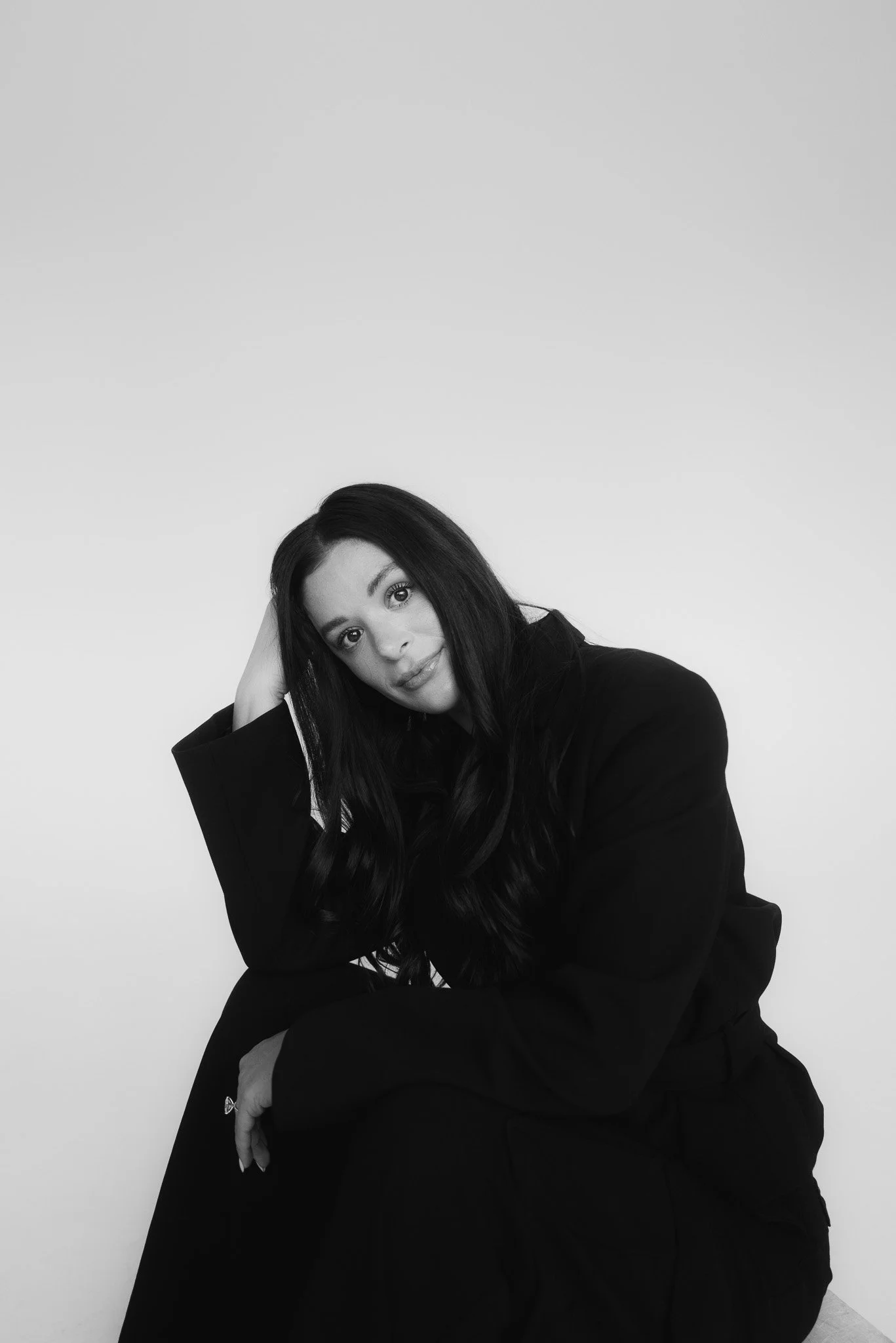 Black and white portrait of a woman with long dark hair, sitting and resting her head on her hand, wearing a dark blazer, against a plain light background.
