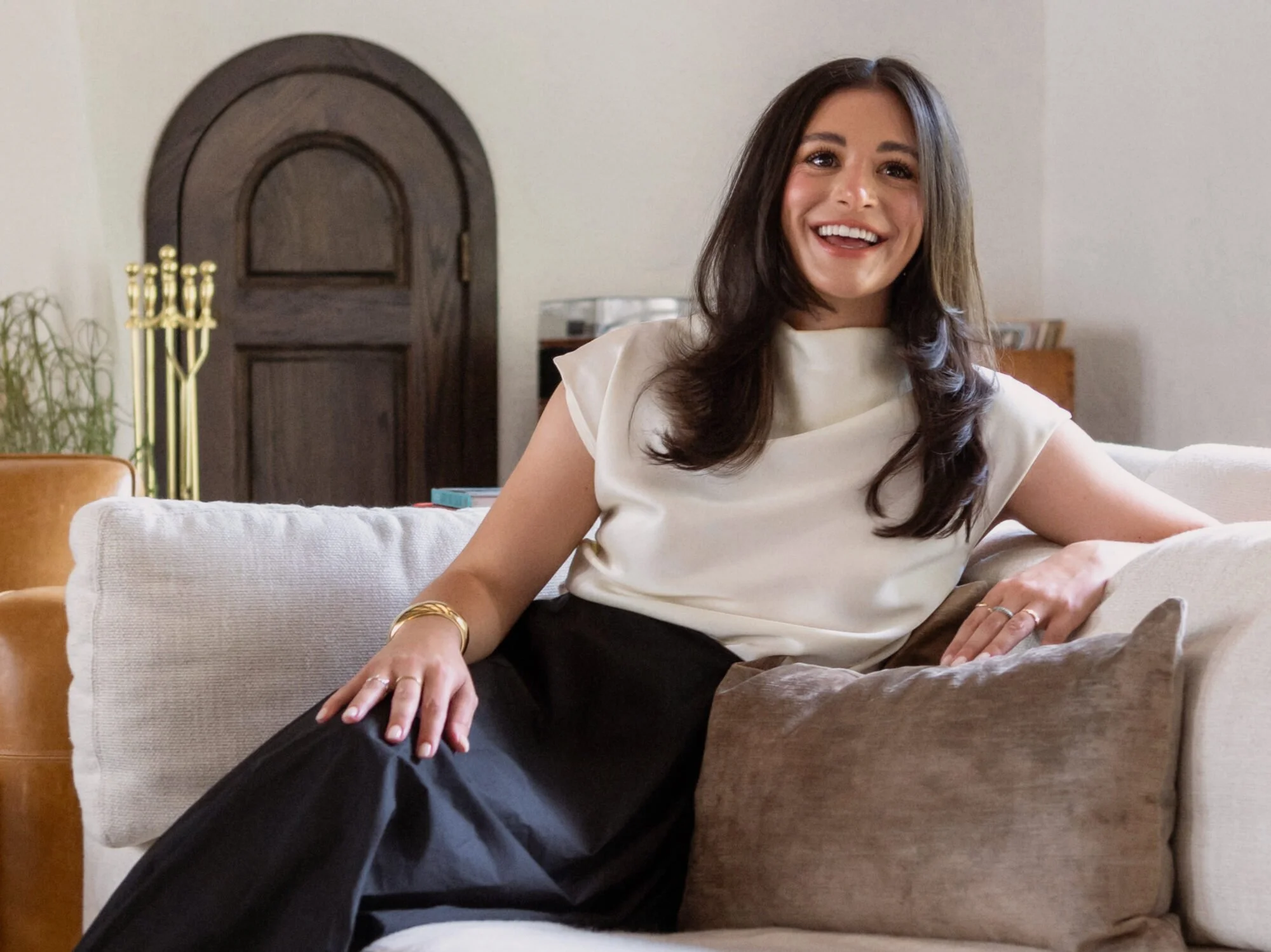 A woman with long dark hair smiling, sitting on a couch in a bright living room, wearing a cream-colored top and black pants, with a wooden door and decorative plants in the background.