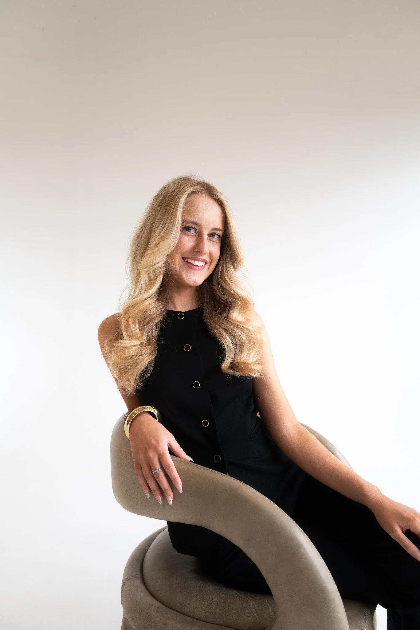 A young woman with long blonde hair, wearing a black sleeveless dress, sitting on a beige modern chair with a rounded backrest, smiling at the camera against a plain white background.