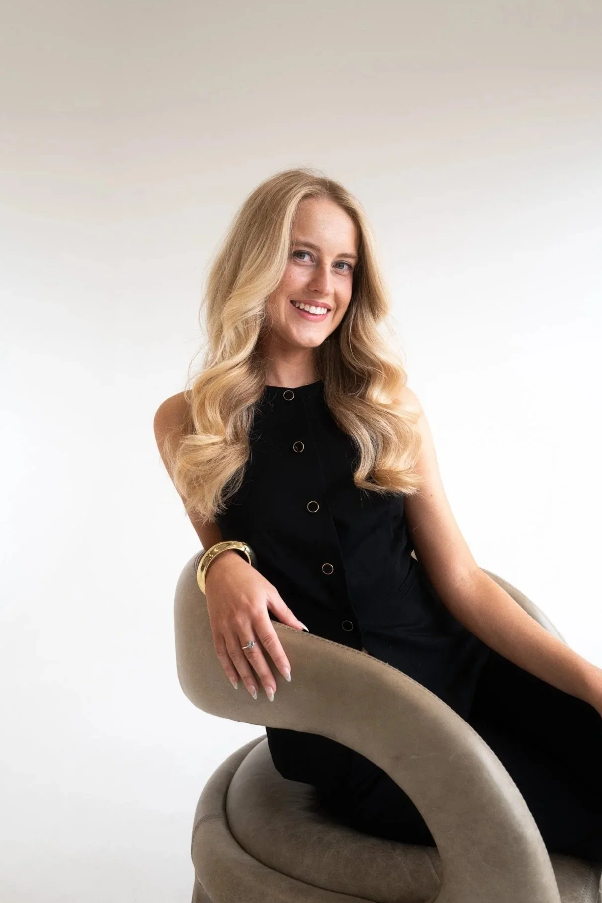A young woman with long blonde wavy hair sitting on a beige modern chair against a plain white background, wearing a sleeveless black dress, gold jewelry, and smiling.