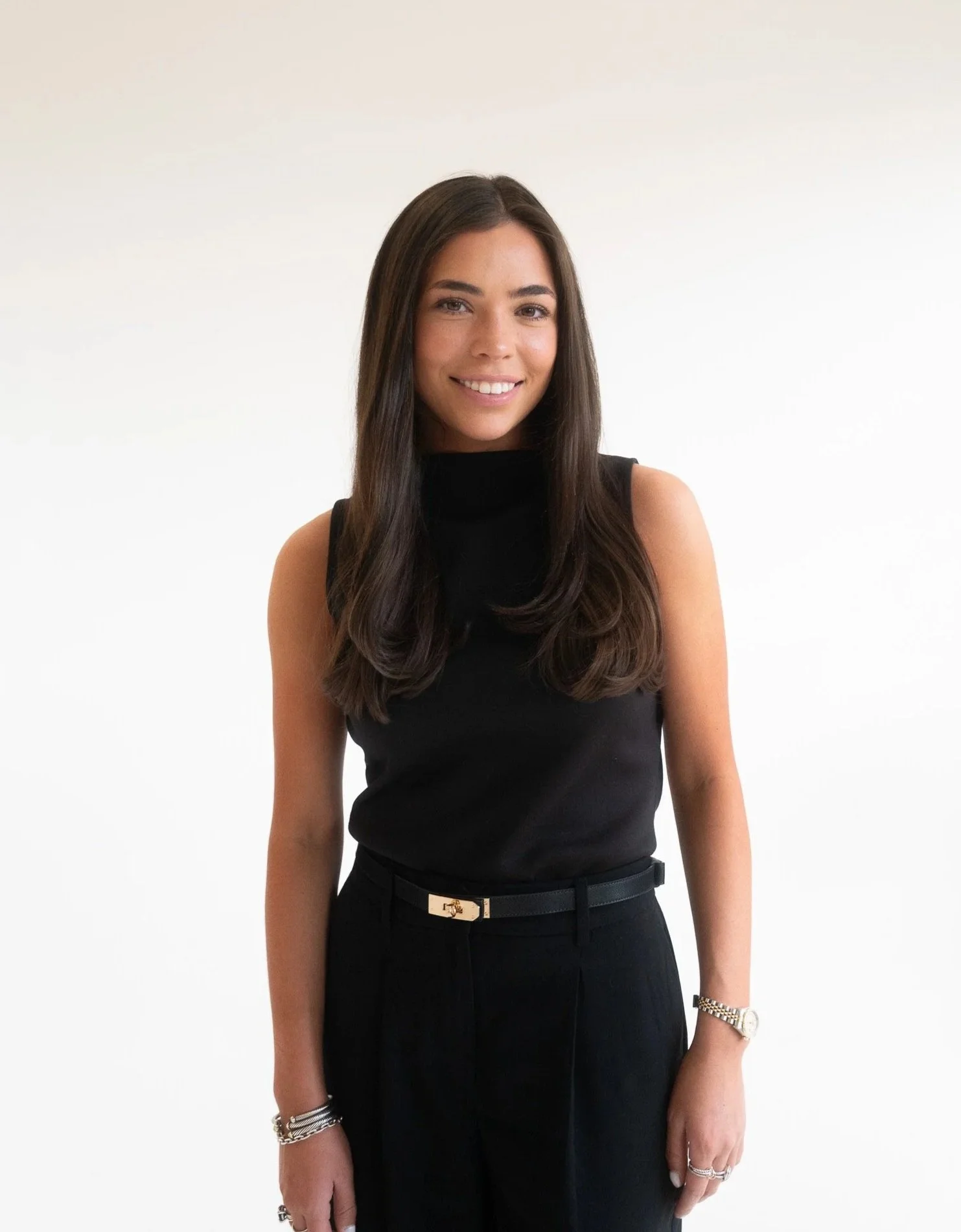 A woman with long dark hair wearing a sleeveless black top, black pants, a black belt with gold accents, a silver watch, and jewelry, standing against a plain white background, smiling at the camera.