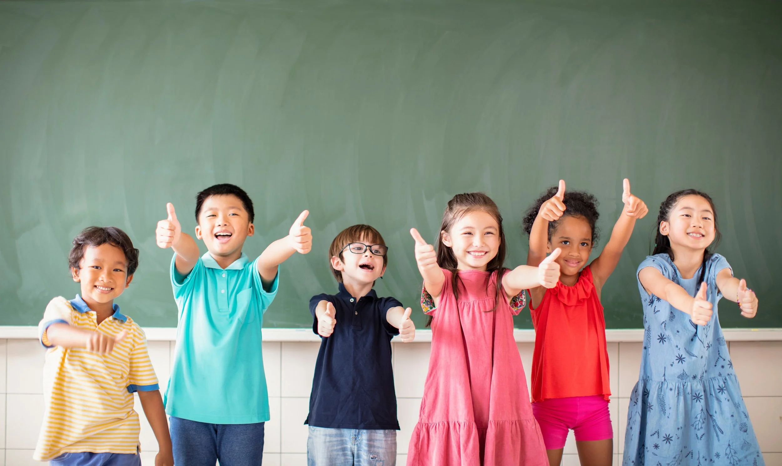Six children standing in front of a chalkboard, smiling and giving thumbs up.