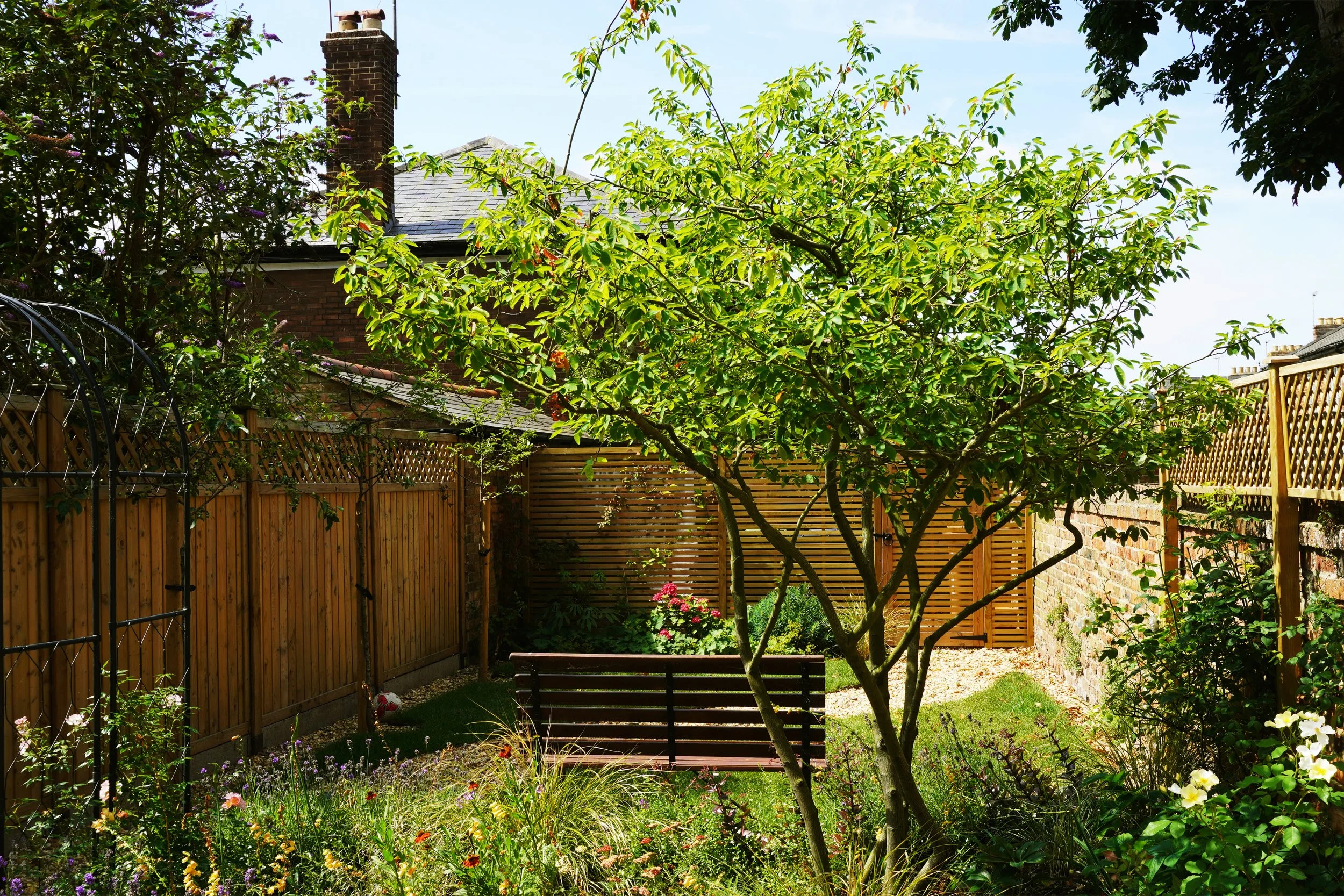 An Oxford city garden design elevation showing an outdoor area with trees, shrubs, a seating bench, a fence, and a person standing on a balcony reading a book. There are also smaller plants and a sun in the sky.