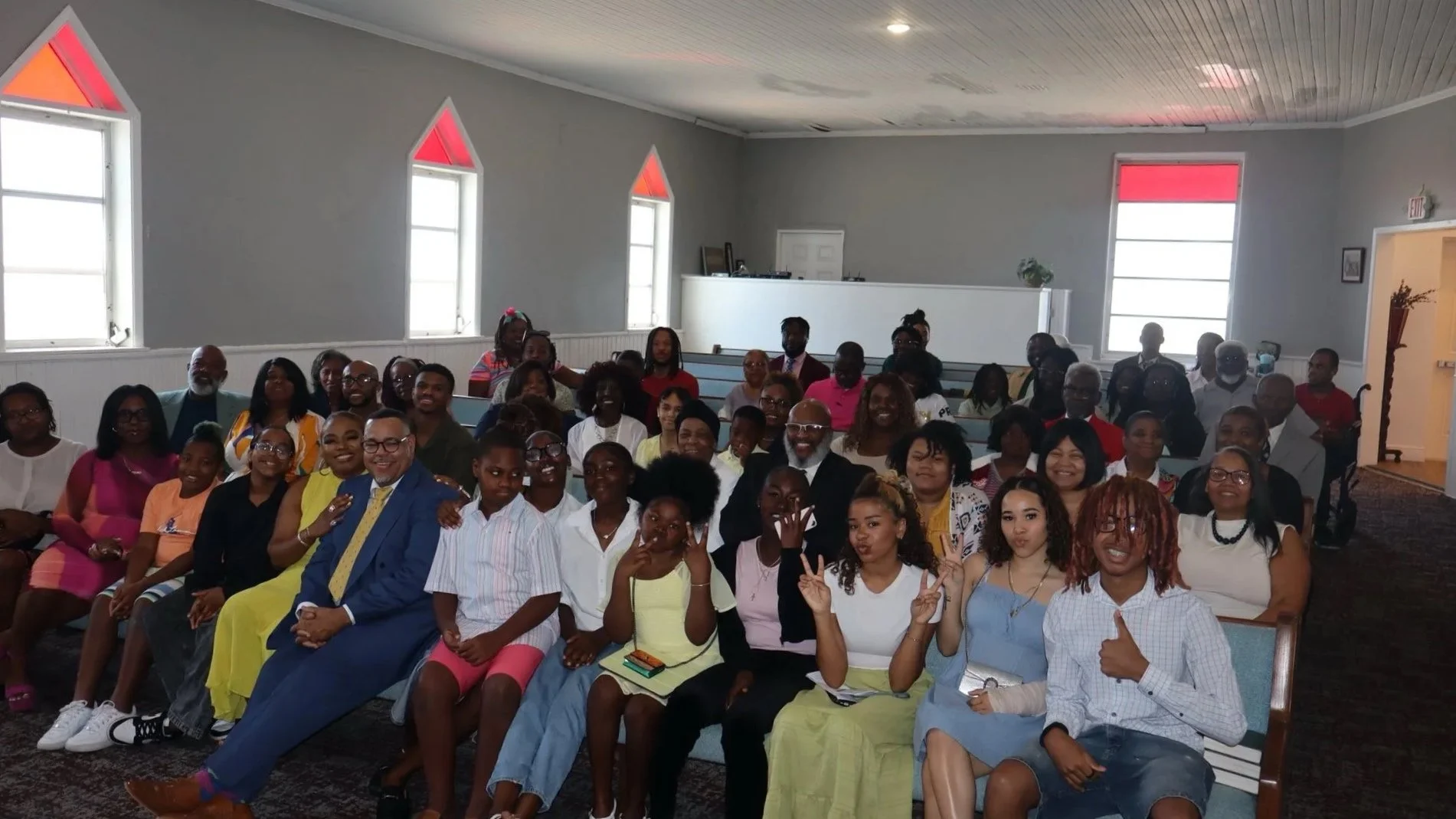 A large group of people gathered in a church or community hall, posing for a group photo, with some making peace signs.