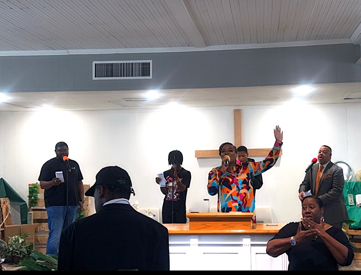 A church service with people singing and speaking at the front, including a woman in a colorful jacket raising her hand, a man in a suit, and children, with a congregation watching.