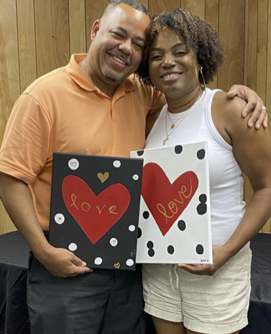 A smiling man and woman holding matching greeting cards with red hearts and the word 'love' written on them, standing in front of a wooden wall.