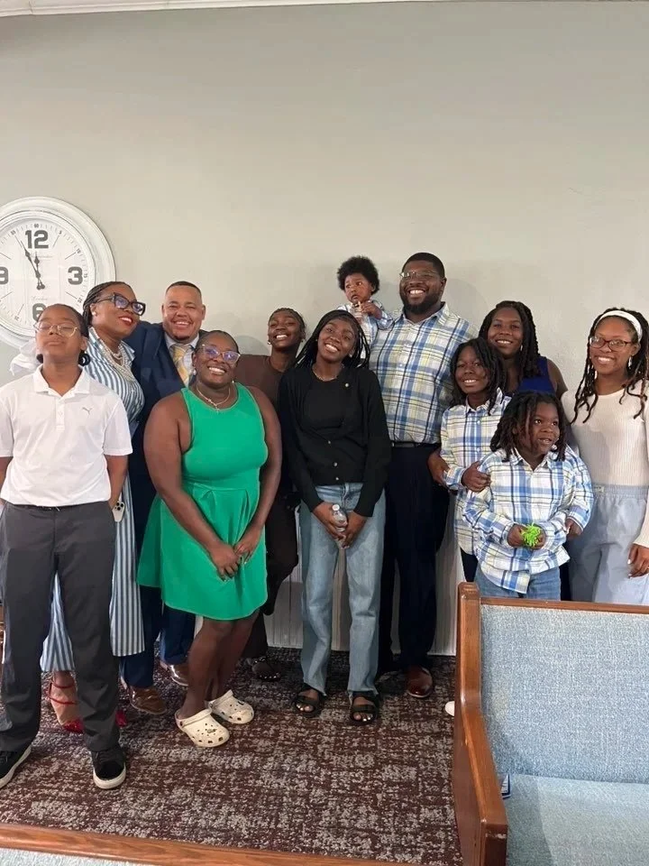 Group of people smiling and standing together indoors, with a clock on the wall behind them.