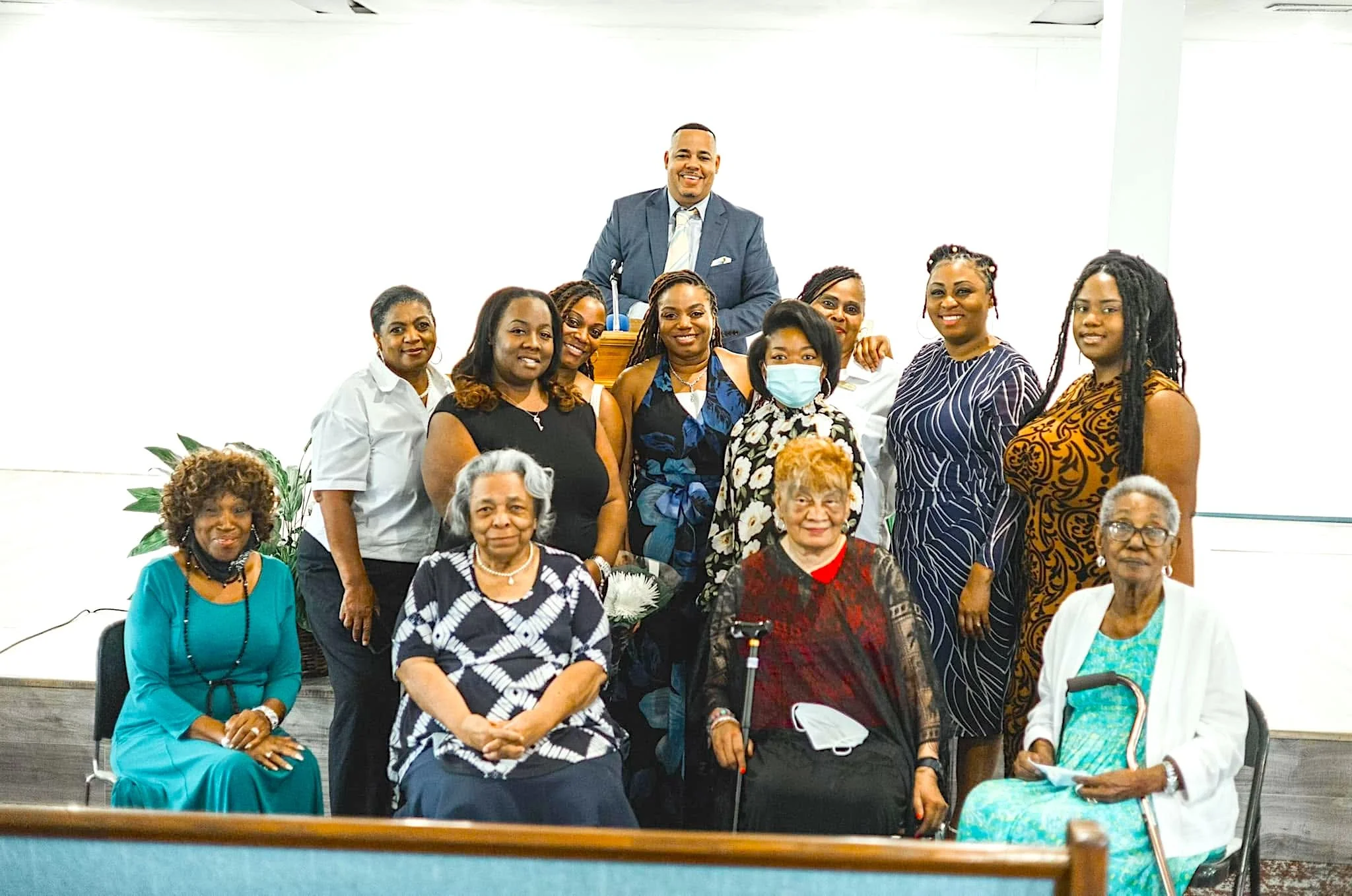 A group of women and one man gathered for a photo in a brightly lit room. The man is standing behind a wooden podium, wearing a suit and smiling. The women are dressed in various colorful and patterned outfits, some seated and some standing. One woman is wearing a face mask, and some are holding flowers or papers.