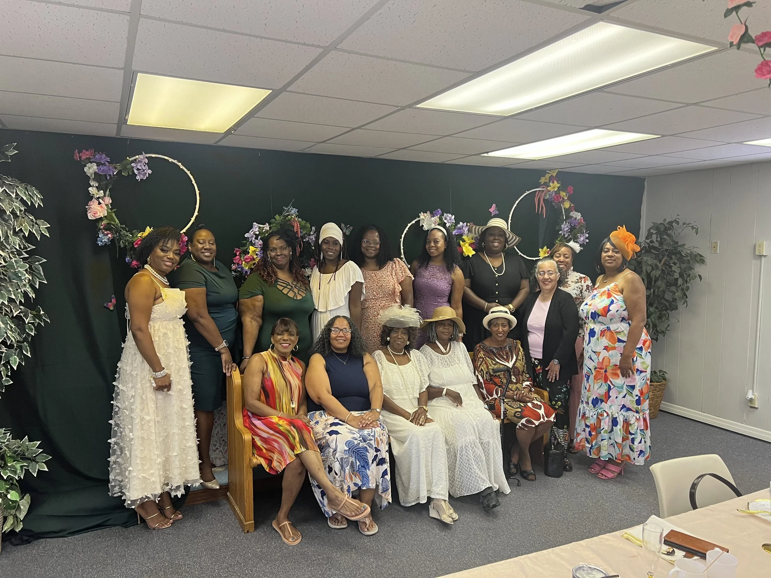 Group of women, mostly wearing colorful dresses, gathered together in a decorated indoor setting with floral arrangements, some wearing hats, smiling for a group photo.