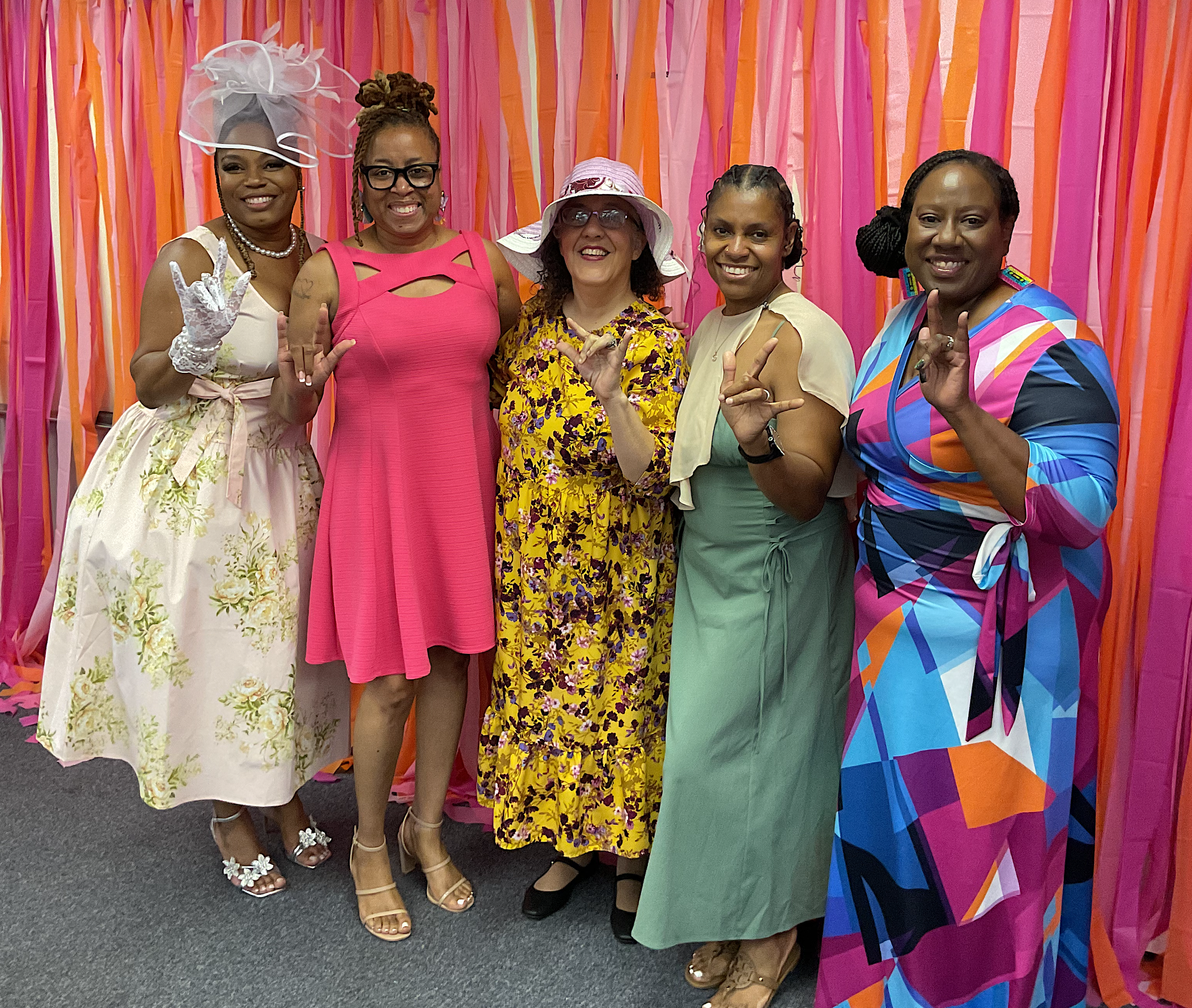 Group of five women standing in front of a pink and orange streamers backdrop, dressed in colorful and floral clothing, posing with peace signs and smiling at the camera.